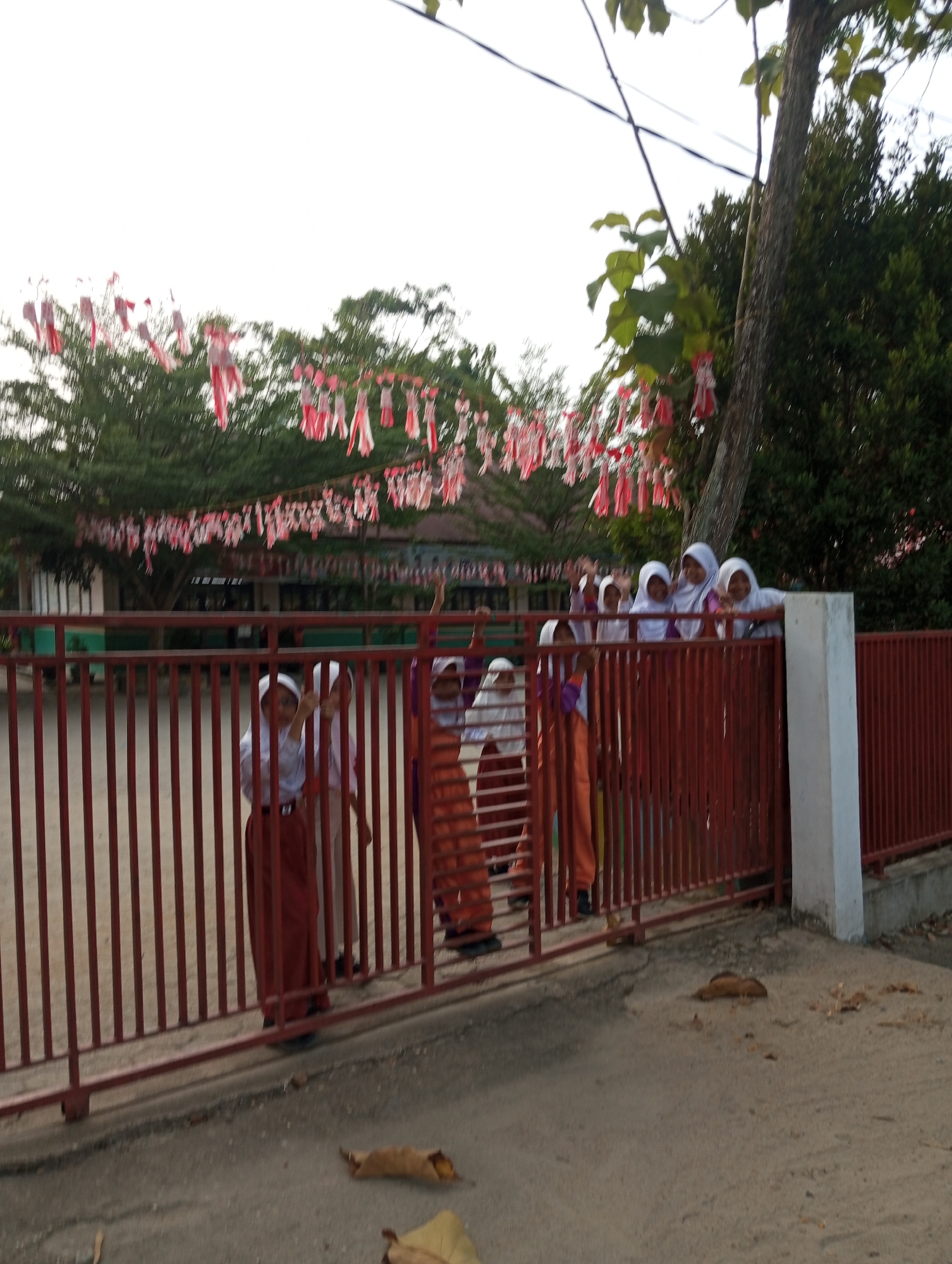 A group of seven young students, all wearing white headscarves and uniforms consisting of white shirts and maroon skirts or pants, are visible behind a red metal fence. The students appear to be standing on the grounds of a school in Pematang Reba, Indonesia, likely during an event given the red and white streamers hanging in the background, possibly for Indonesian Independence Day. The students are waving and looking towards the camera. The fence is the dominant foreground element, with a building and lush green trees visible behind it. The ground in front of the fence is paved, with a few fallen leaves scattered on it.