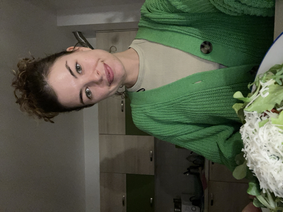 A young adult female is depicted in a kitchen setting located in Boreti, Montenegro. She has light skin, light-colored eyes, and dark, curly hair styled in an updo. She is wearing a light beige ribbed t-shirt under an open, bright green knitted cardigan, secured by one visible brown speckled button. The subject is looking directly at the camera with a slight smile. In the immediate foreground, a white bowl with a blue rim contains a salad composed of green leafy vegetables, heavily topped with a grated white substance, consistent with cheese. The background features a kitchen area with light wooden upper cabinets and darker green lower cabinets. A white appliance, possibly a microwave, and various kitchen utensils are visible on the countertop. The environment is illuminated by interior lighting.