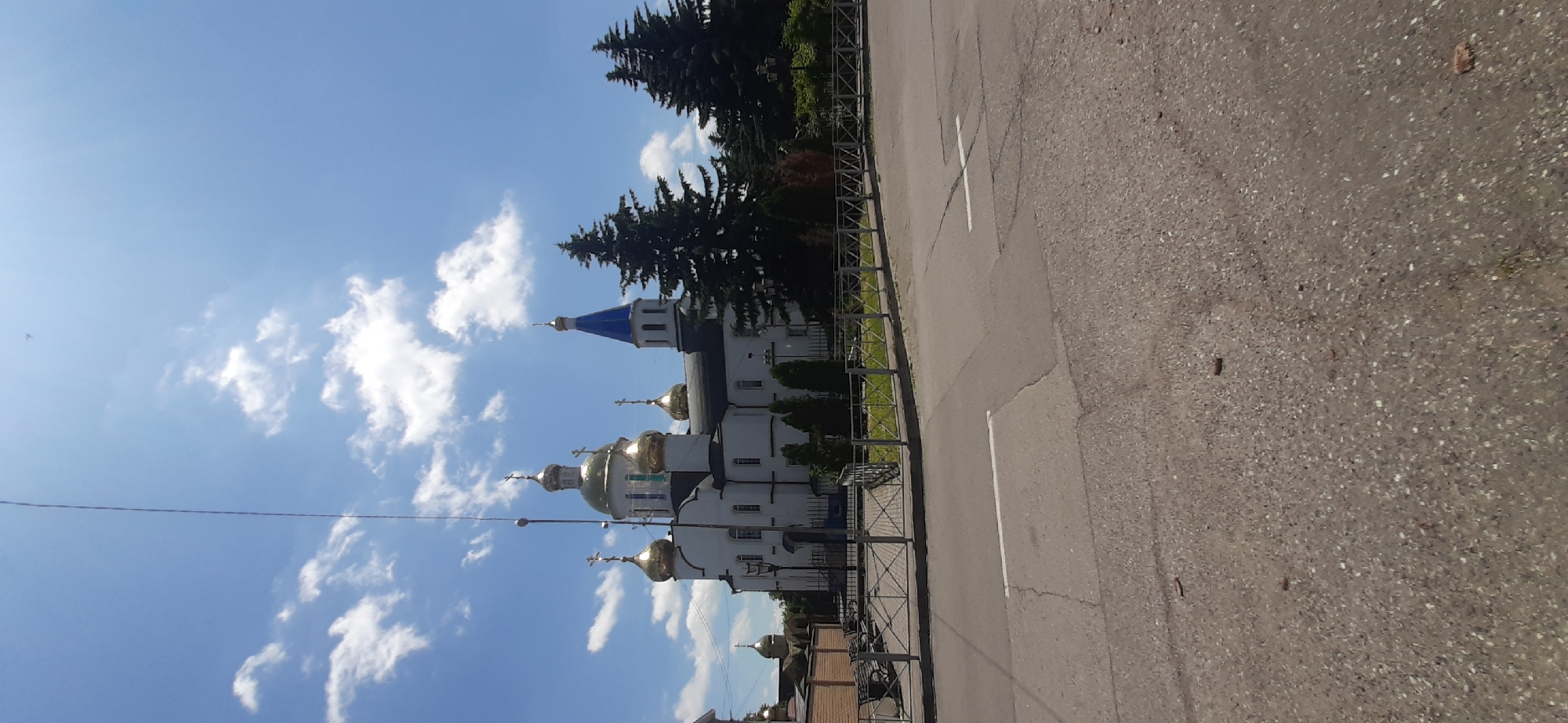 The image captures a view of a white church with multiple golden domes, characteristic of Orthodox Christian architecture. A single blue-topped tower stands out among the golden domes. The church is surrounded by a fence, and behind it are several large evergreen trees. The sky is a clear blue with scattered white clouds. In the foreground, a paved road with white markings is visible, suggesting an urban or suburban setting. There are no people or animals apparent in the frame. The overall scene depicts a religious building under a bright sky on what appears to be a clear day. The context provided about Yessentukskaya, Russia, suggests this is a church located in that region.