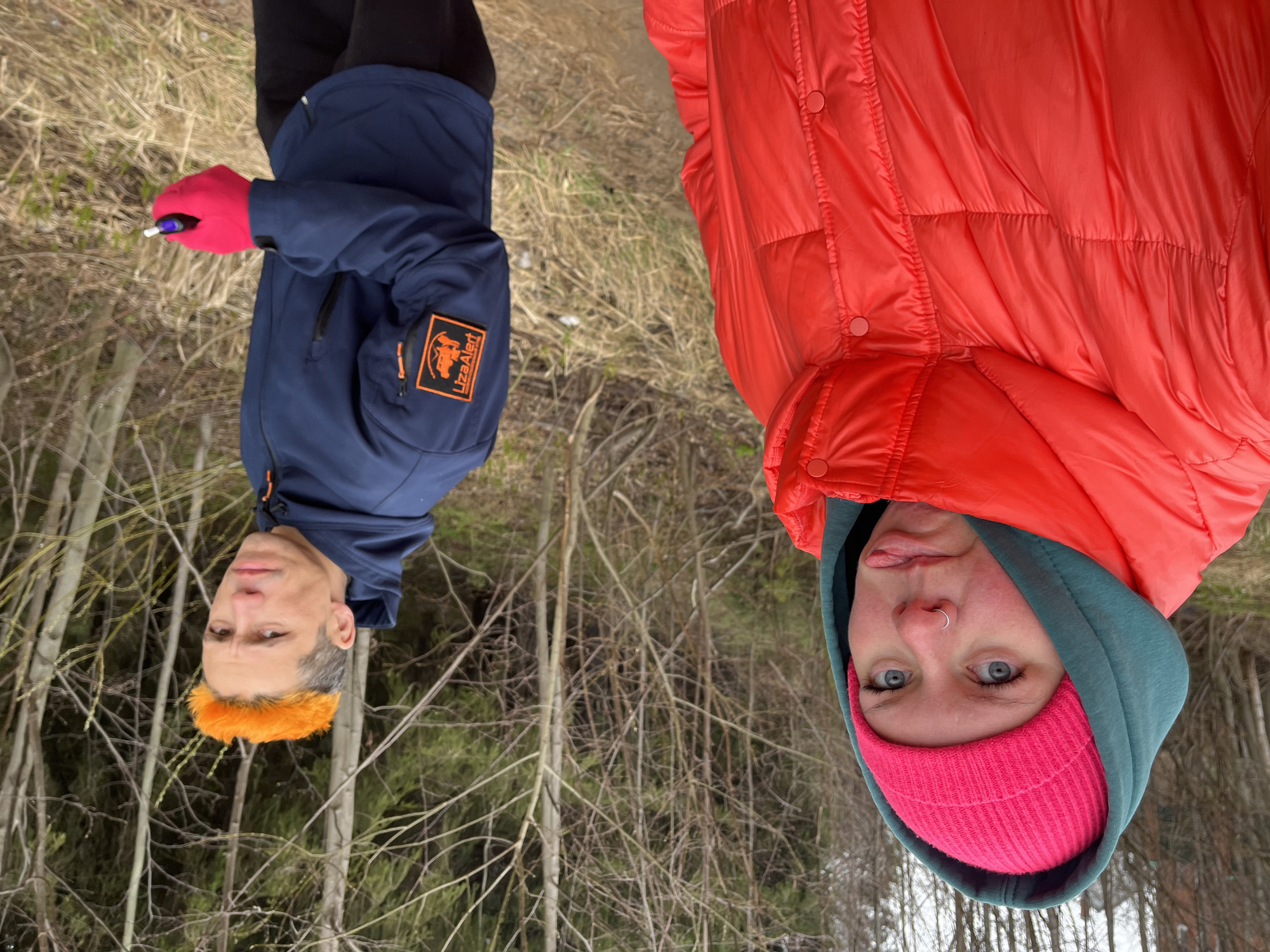 Two people are visible outdoors in a wooded area, likely in Chaikovsky, Russia, as indicated by the "LizaAlert" logo on the jacket of the person on the left. The person on the left, a man with short, grey hair and what appears to be a bright orange hairpiece, is wearing a blue jacket with an orange patch that reads "LizaAlert" and red gloves. He is holding a small object in his gloved hand. The person on the right, a woman, is wearing a bright pink beanie, a teal hoodie, and a bright orange puffer jacket. The background consists of bare trees, brown grass, and snow-covered ground.