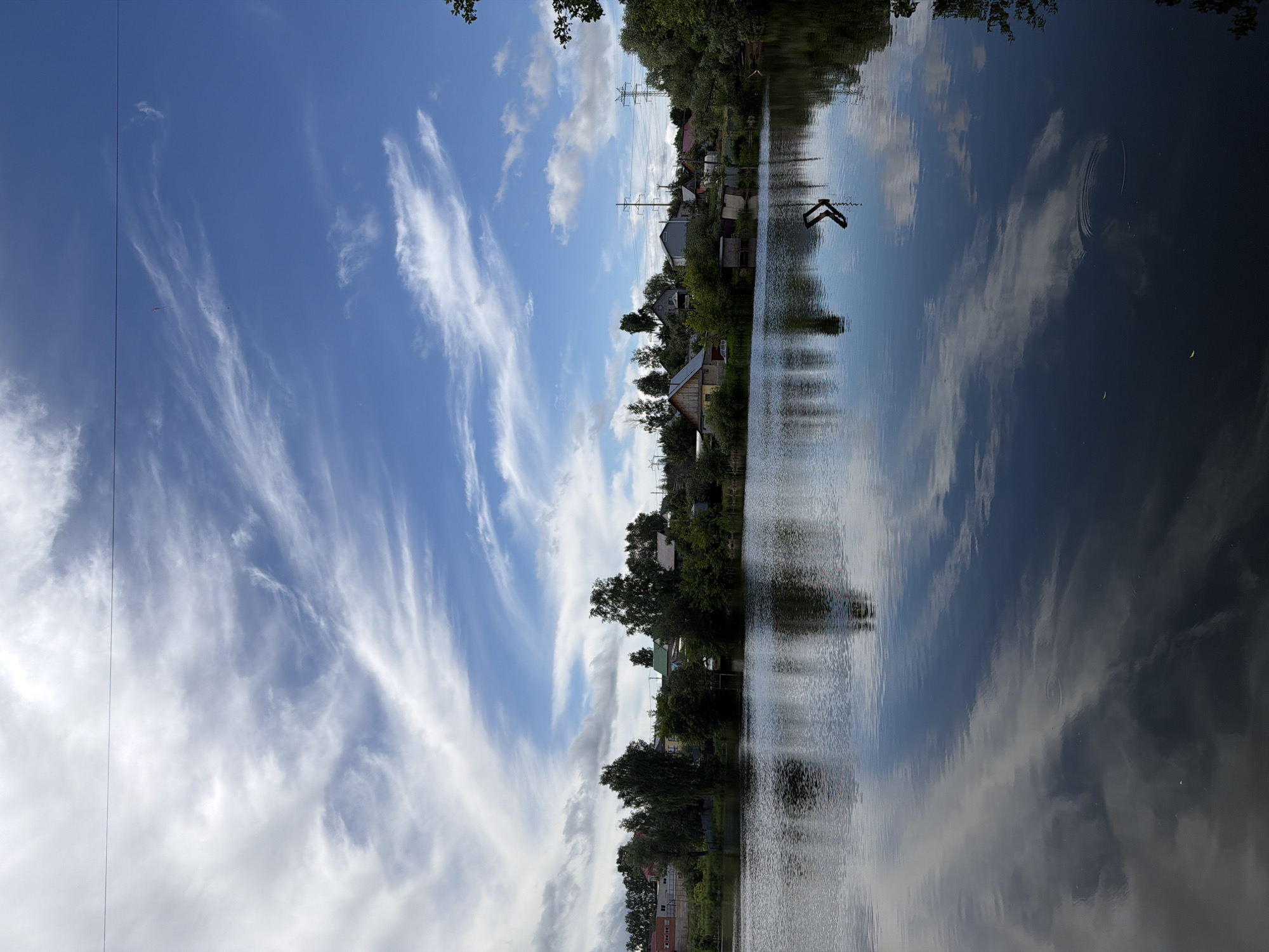 The image depicts a serene lakeside scene in Chaikovsky, Russia. The foreground is dominated by a calm body of water, its surface reflecting the bright blue sky adorned with wispy white clouds. The reflections of trees and structures on the far bank are clearly visible on the water.
The middle ground features a lush green shoreline populated by various trees and a cluster of residential buildings, indicative of a village or suburban area. Among the structures, one can discern houses with pitched roofs and possibly some utility poles. A hint of a road or path may be present near the buildings.
In the background, a vibrant blue sky with scattered cirrus clouds creates a dynamic contrast with the tranquil water. The overall impression is one of a peaceful, natural setting on a clear day. There are no people or animals visible in the image. The scene appears to be a typical summer or early autumn day in a rural or semi-rural part of Chaikovsky.