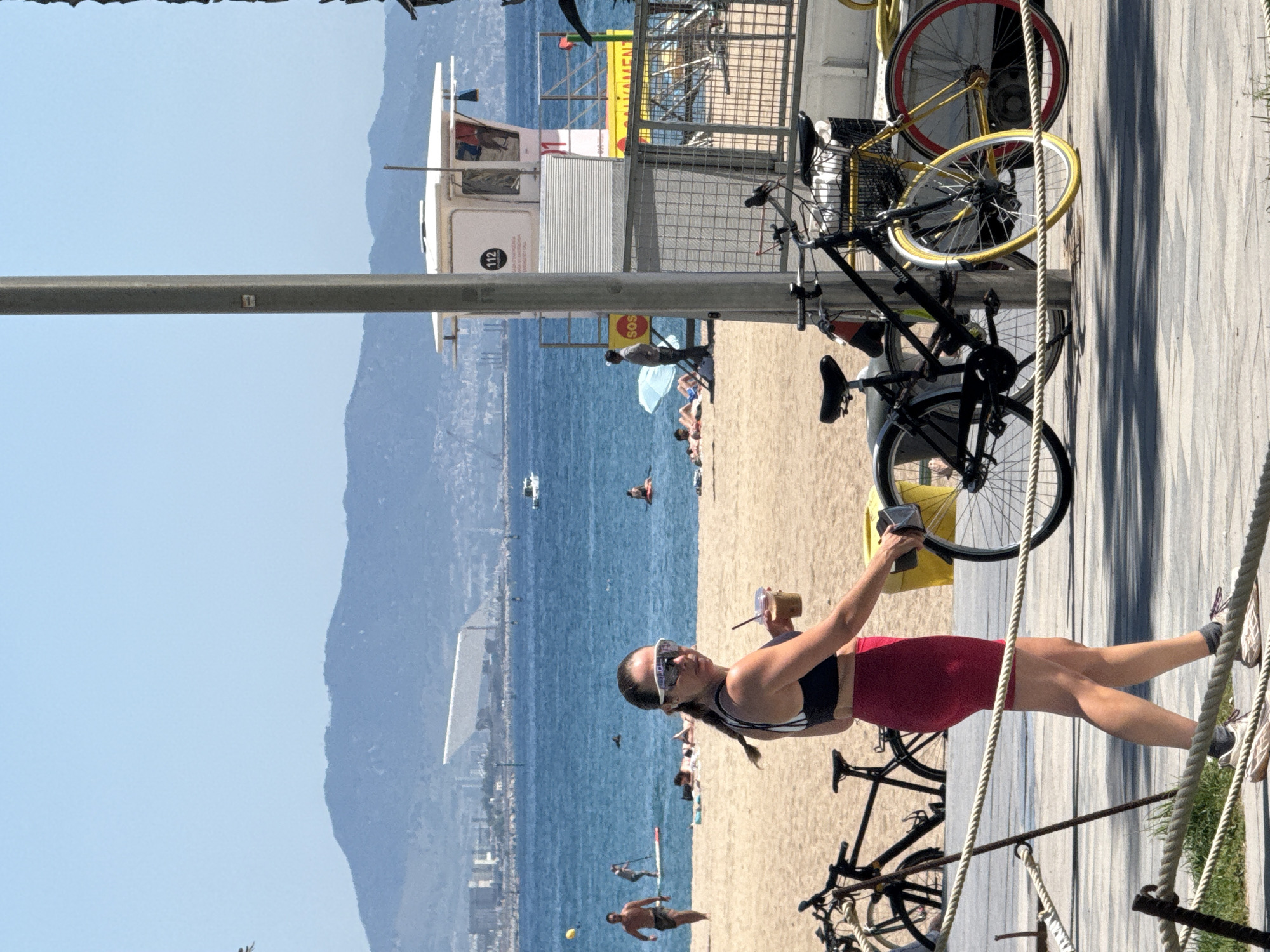In Barcelona, Spain, on a sunny day at the beach, a woman with a ponytail is standing on a wooden boardwalk, holding a drink with a straw. She is wearing a black sports bra and red shorts, with white athletic sunglasses. In the background, the beach is populated with people sunbathing and swimming in the blue ocean. A lifeguard tower stands prominently on the beach, with a few bicycles parked on the boardwalk. In the distance, the city skyline and mountains are visible.