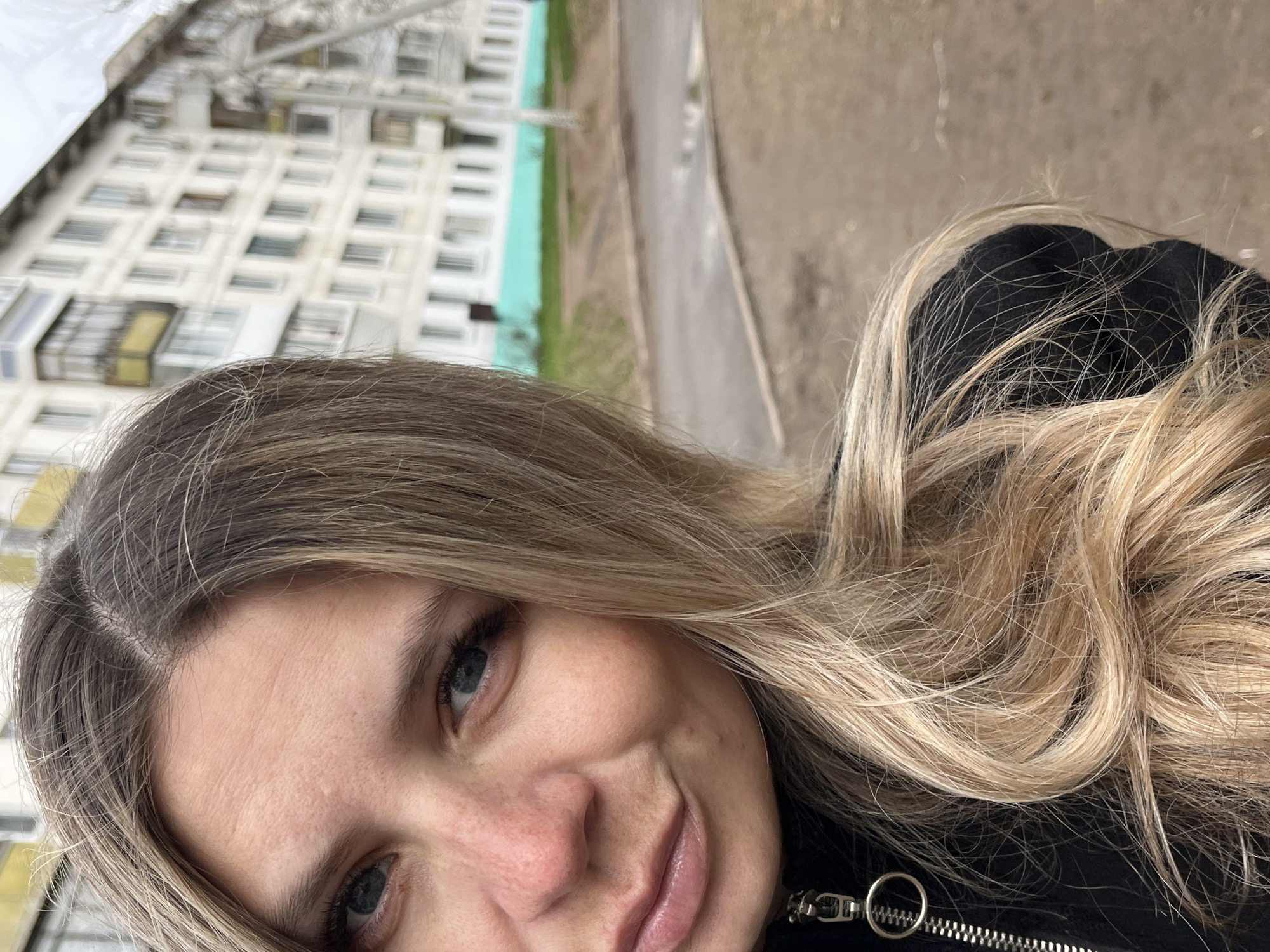 A close-up selfie of a woman with blonde hair and blue eyes. She is smiling slightly. The background shows an apartment building with multiple windows and a patch of green space. A section of road is also visible. The apartment building architecture and overall scene suggest a location in Chaikovsky, Russia. The woman wears a dark jacket.
