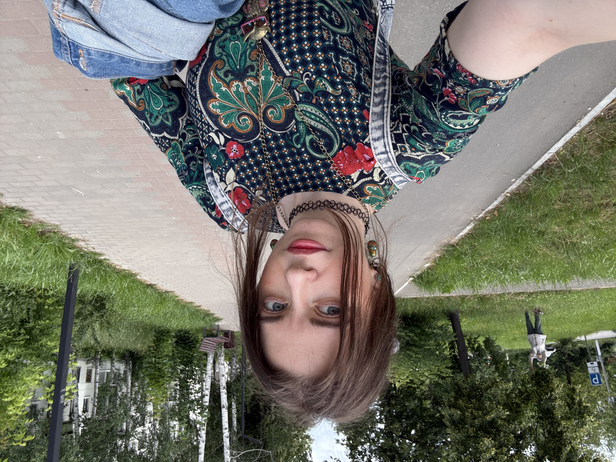 A young woman with light brown hair is taking a selfie outdoors on a paved path. She is wearing a dark, patterned dress with red and green floral motifs, a black choker necklace, and a long pendant necklace with an owl charm. She is also carrying a light blue denim jacket. Behind her, the path continues, bordered by green grass and trees. In the distance, there are birch trees, a park bench, a lamppost, and a white building. A person is visible in the background, standing in the grass. The weather appears overcast.
