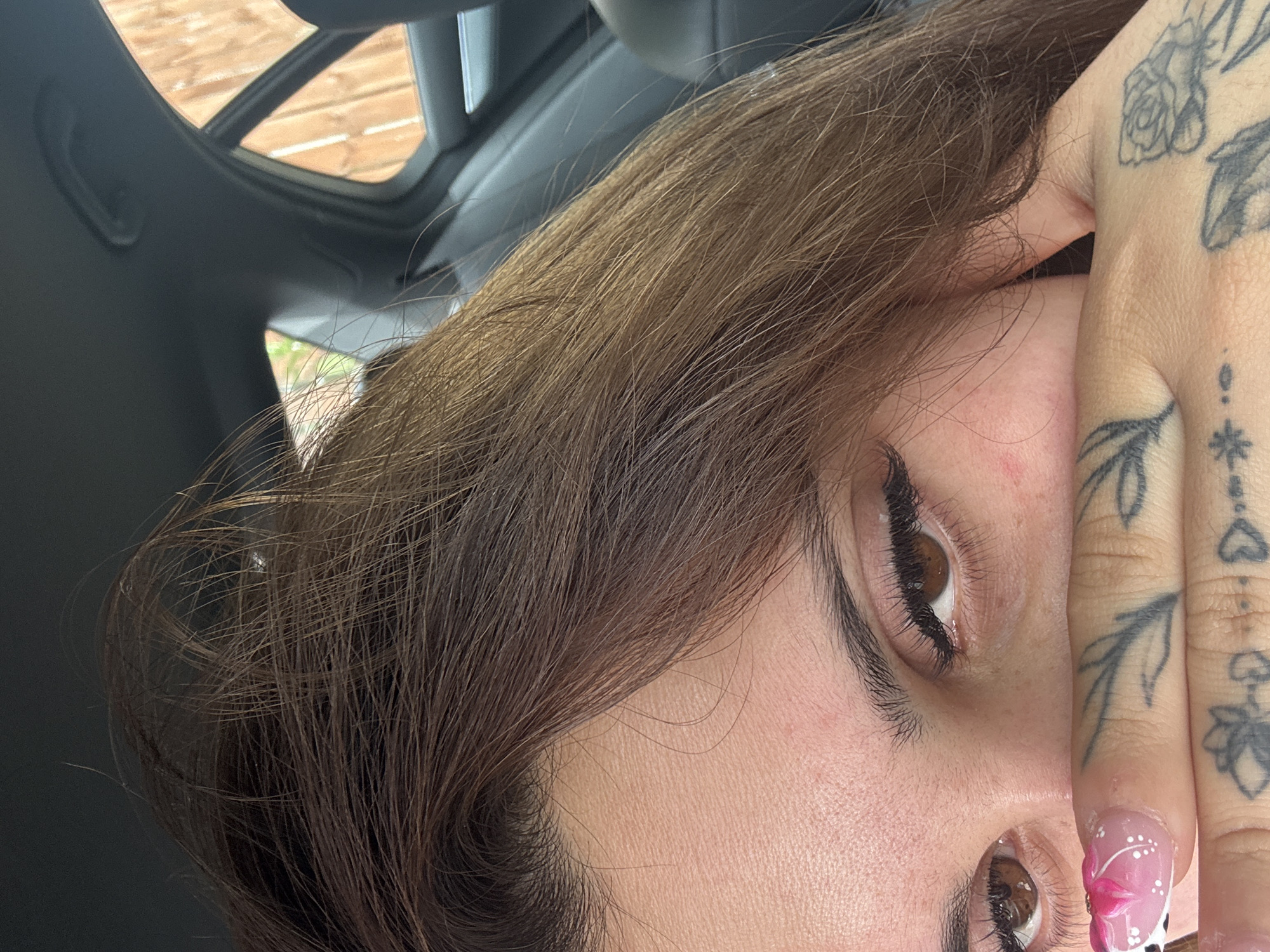 The image shows a close-up of a person's face, with their hand covering their mouth. The person has brown hair and brown eyes, and is wearing eyeliner and false eyelashes. Their fingernails are decorated with pink and white patterns. There are tattoos visible on their hand. The background suggests the person is inside a car, with a view of a wooden fence through the car window. The location of Dungannon, United Kingdom, is not discernible from the image itself.