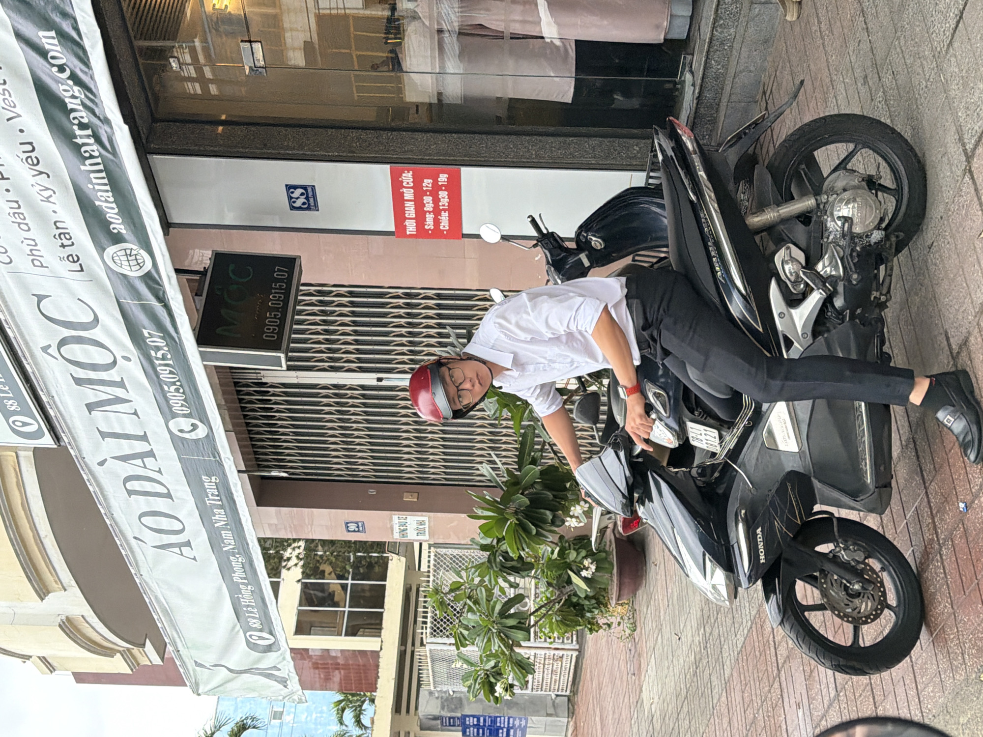The image captures a street scene in Nha Trang, Vietnam, featuring a person on a black Honda scooter. The scooter is parked on a tiled sidewalk in front of a shop with a large banner. The banner, in Vietnamese, reads "ÁO DÀI MỘC" and displays the address "88 Lê Hồng Phong, Nam Nha Trang" and a phone number "0905.0915.07". Above this, another sign indicates a website "aodainhatrang.com".
The person on the scooter is wearing a white collared shirt, black trousers, black dress shoes, and a red helmet. They appear to be looking towards the viewer. The scooter's license plate is partially visible.
To the right of the scooter, the shop's glass door reflects some interior elements. A red sign on the door lists "THỜI GIAN MỞ CỬA:" (Opening Hours) with times for morning and afternoon. Further to the right, mannequins or displays can be seen inside the shop.
The background includes architectural elements of buildings, a potted plant with flowers, and other street furniture. The lighting suggests it is daytime.