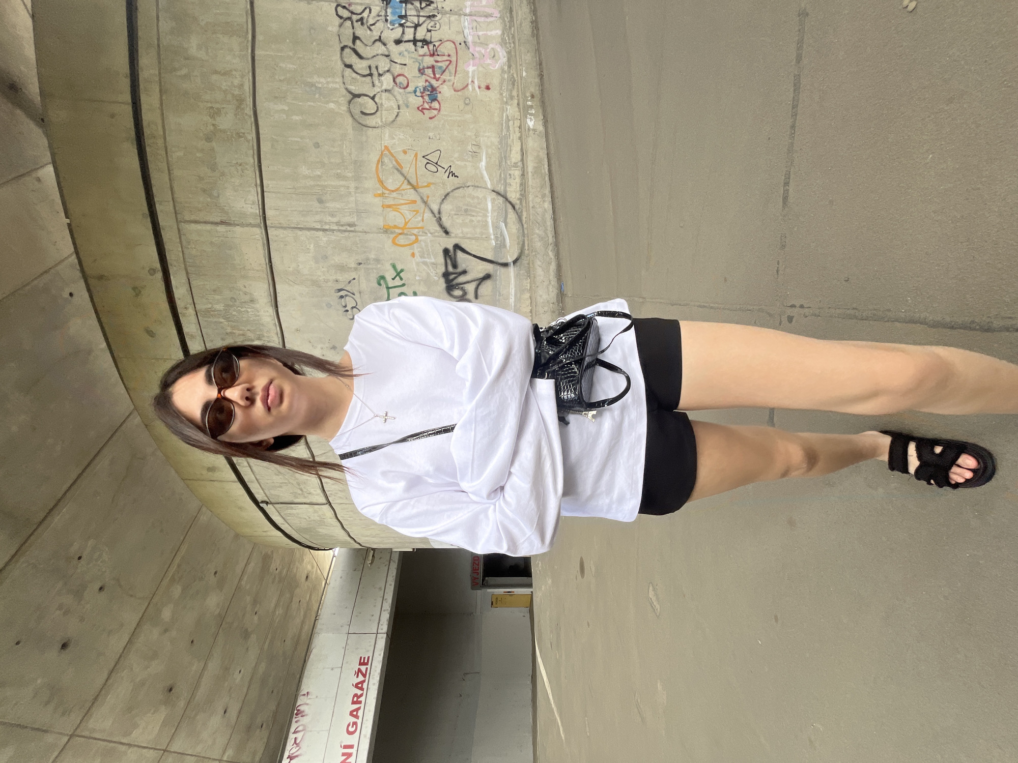 A young woman stands facing the camera with her arms crossed, wearing a white long-sleeved shirt, black shorts, and black sandals. She has on sunglasses and a small black crossbody bag with a textured pattern. Behind her is a concrete structure with graffiti on it, and to her left, a building with a sign that reads "NÍ GARÁŽE" (Czech for "Garage"). The location is the underpass of the D5 highway in Pilsen, Czechia, near the Pilsen University Hospital. The lighting suggests it is daytime.