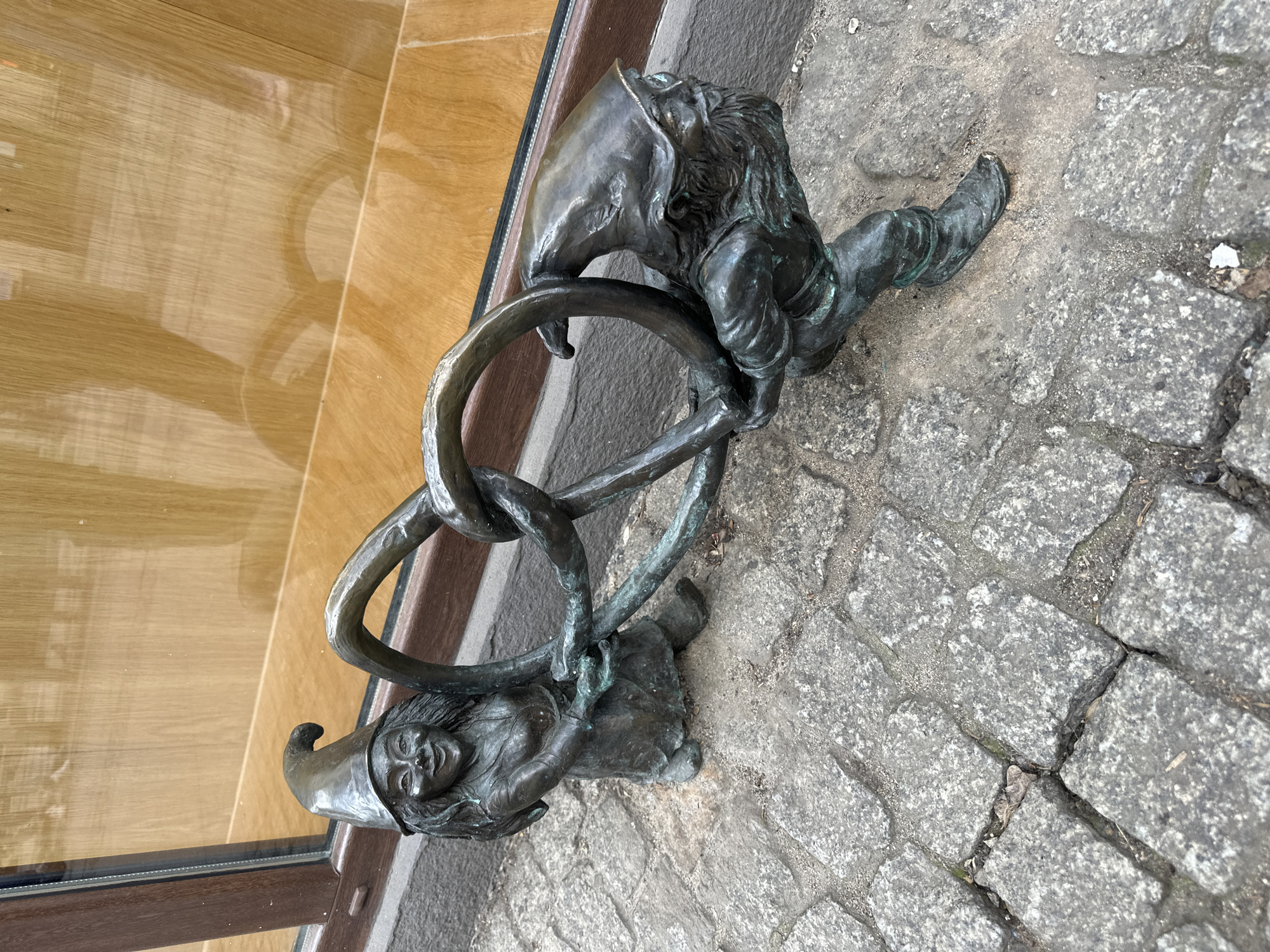 A statue of two Wrocław dwarves, or "krasnale," climbing through interlocked rings, is located on the street in Wrocław, Poland. One dwarf wears a pointed hat and smiles upwards. The other has long hair and is attempting to hoist itself through the rings. The statue sits on a cobblestone surface, and part of a building façade is visible in the background.
