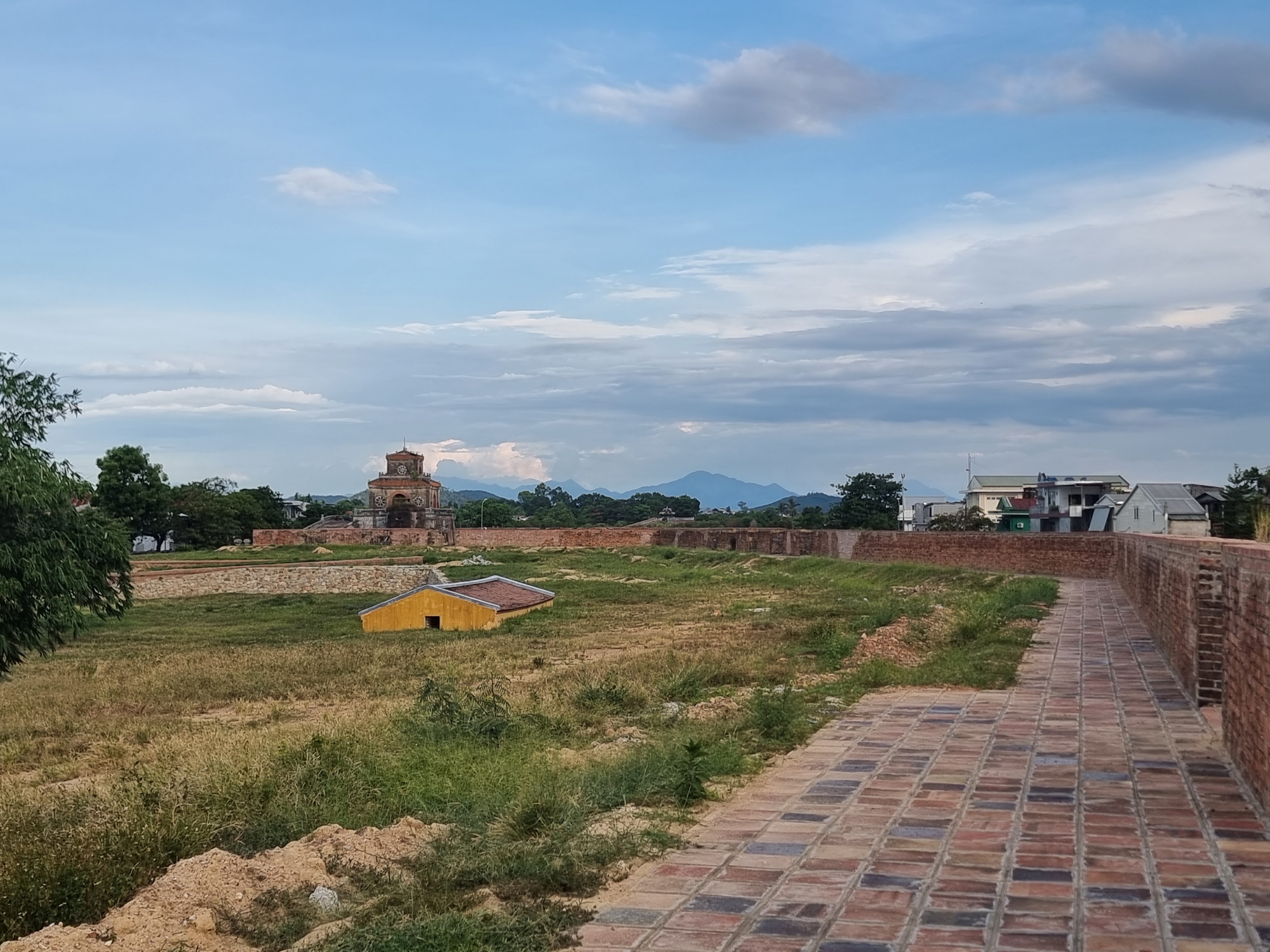 The image depicts a view of the Imperial City of Huế, located in Phú Xuân Ward, Vietnam. In the foreground, a brick pathway runs along the top of a brick wall. To the left of the pathway is a grassy field with dry grass and some green vegetation. A small, yellow, unoccupied building with an orange roof is situated in the field. Further in the background, an ornate, multi-tiered tower-like structure with a circular design on top is visible, constructed from brick. Behind this structure, there are trees, other brick walls, and a cluster of buildings that appear to be modern houses. In the far distance, a range of mountains under a cloudy sky can be seen. The overall scene suggests a historical site with elements of nature and modern development.
