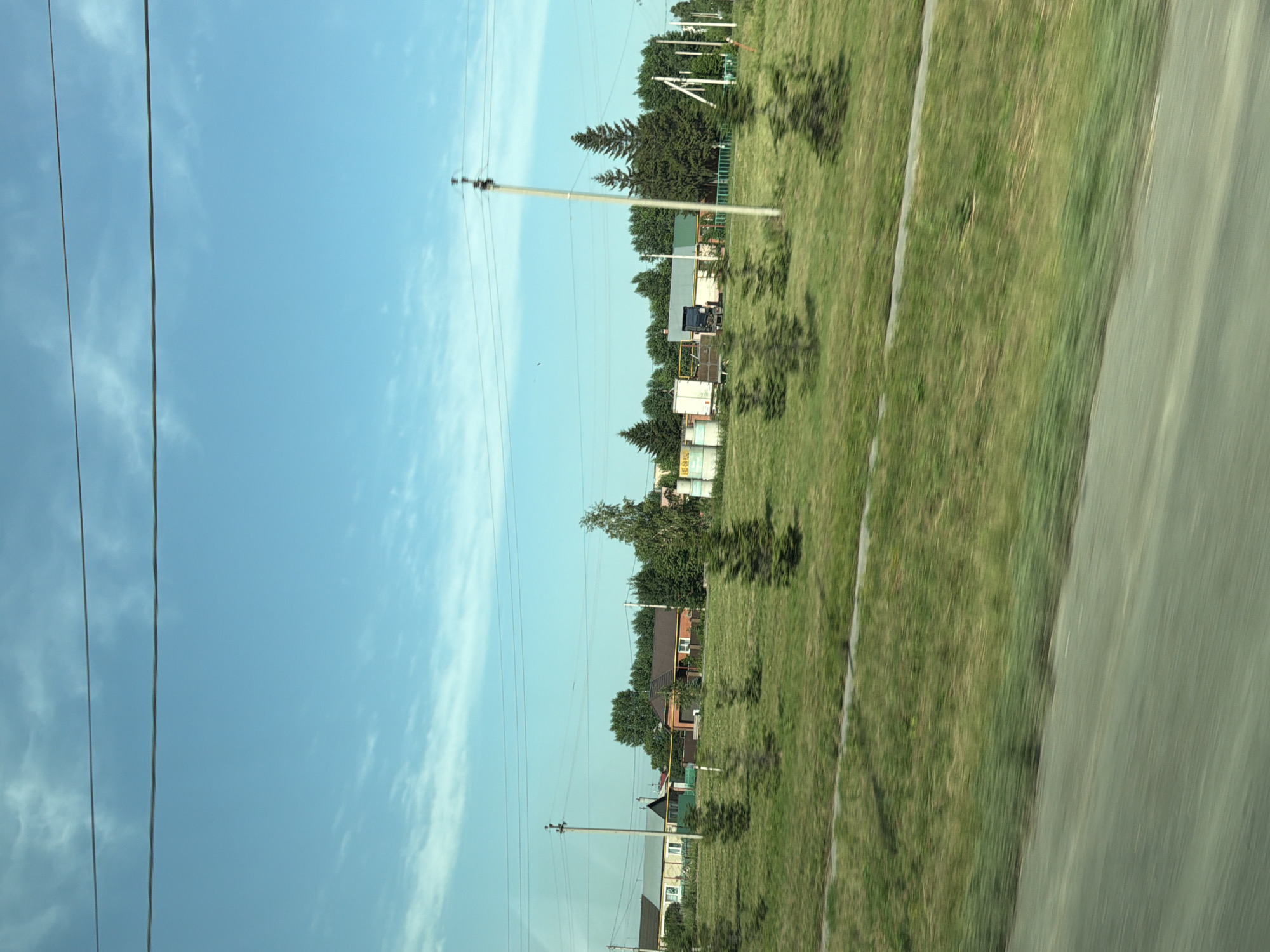 This image captures a rural landscape on a bright, clear day, likely in Evsino, Russia. The scene is dominated by a vast expanse of bright blue sky with scattered wispy clouds. Two parallel power lines stretch horizontally across the upper portion of the frame.
In the middle ground, a row of modest, single-story houses with pitched roofs is visible, interspersed with mature trees and evergreen shrubs. Some houses appear to have brick or painted exteriors. A paved road runs in front of the houses, bordered by a grassy verge. Behind the houses, a dense line of tall trees forms a natural barrier.
To the right, several large, cylindrical concrete structures are piled together, suggestive of an industrial or construction site. A truck is parked near these structures. A tall, utility pole with crossbars stands prominently in the middle of the scene, also carrying power lines. The foreground shows a blurred grassy field, indicating movement, possibly from a vehicle. The overall impression is of a peaceful, sunlit day in a rural settlement.