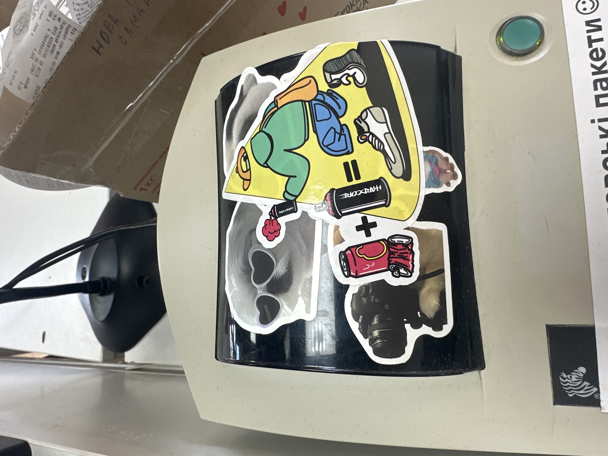 The image shows a close-up view of a beige and black electronic device, possibly a scale or a label printer, located on a counter. The device is adorned with several stickers. A prominent sticker features a dog wearing heart-shaped sunglasses. Another sticker depicts a camera, specifically a Canon EOS 6D. There's also a sticker of a red can with a yellow circular design and another sticker showing a graphic illustration of a person in a green hoodie and blue jeans, holding a spray can, with an equal sign connecting it to a sneaker and a face. A small sticker of a piglet is visible below the illustration. In the background, a brown cardboard box with some writing in Ukrainian is visible, along with part of a black lamp. A small green illuminated button is present on the right side of the device. The overall setting appears to be an indoor commercial environment, likely a post office or a retail store, in Khmilnyk, Ukraine, given the text on the box.