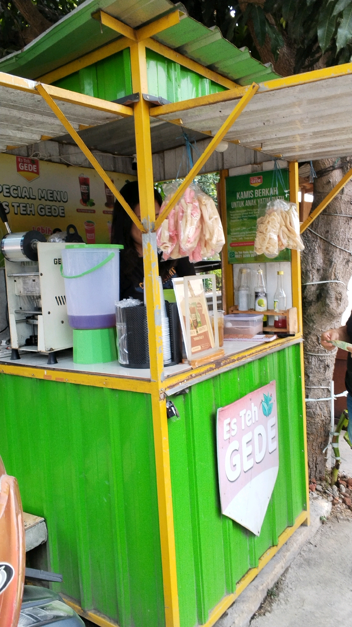 The image depicts an outdoor food stall, identified as "Es Teh GEDE," located in Banjaragung, Indonesia. The stall features a bright green corrugated metal base with yellow trim and a yellow metal frame supporting a green metal roof. Various items are displayed on the counter, including a juice dispenser, buckets, a stack of cups, bags of snacks, and bottles of syrup. A person, partially visible, appears to be operating the stall. A sign in the foreground reads "Es Teh" with a leaf graphic and the word "GEDE" below it. The stall is situated next to a tree, and the ground is a mix of concrete and dirt. The scene suggests a typical street food vendor environment in Indonesia.