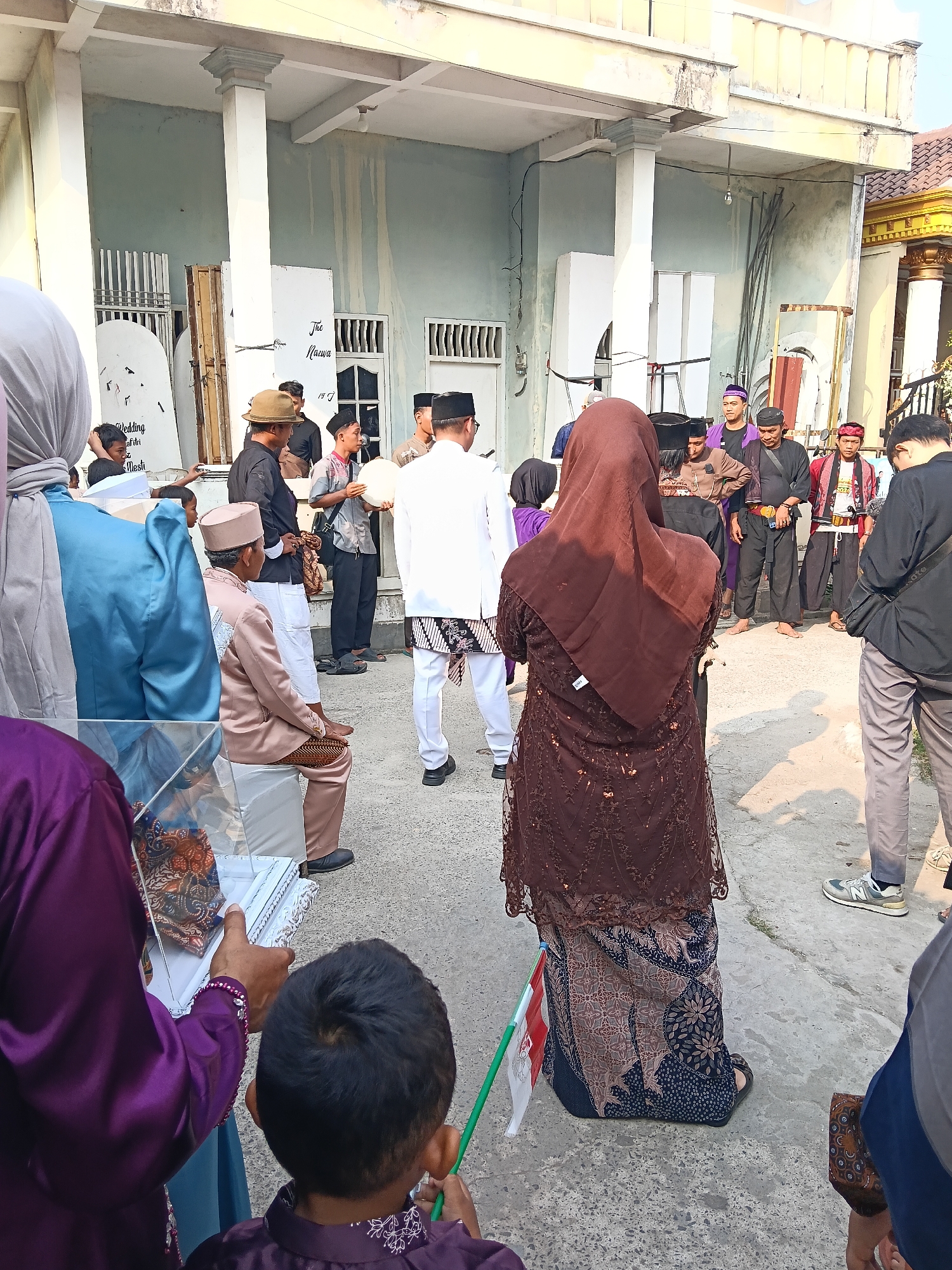 This photograph captures an outdoor gathering in Tegal Angus, Indonesia. The scene appears to be in front of a building with a light blue, weathered facade and white columns. Several individuals are present, many dressed in traditional Indonesian attire.
In the foreground, a person in a purple outfit holds a white box with decorative trim. To their right, a young boy with dark hair looks on, holding a green stick. Behind them, a woman in a brown, patterned hijab and a long, dark brown dress with intricate embellishments stands facing away from the camera. She is holding a small Indonesian flag attached to a green pole.
A group of men are positioned in the mid-ground. One man wears a tan hat and a dark shirt, facing left. Another man, in a light brown traditional outfit, sits on a white chair. Several other men are dressed in various tunics, some with head coverings, suggesting a cultural or ceremonial event. One man in a gray shirt holds a white circular object, possibly a tambourine. Another man wears a dark shirt and pants, with a red sash.
In the background, there are large white boards with writing on them, one of which appears to say "The Nuansa". There are also barred windows and metal structures visible on the building. The ground is paved with concrete. The overall impression is of a community event taking place in a residential or public outdoor space.