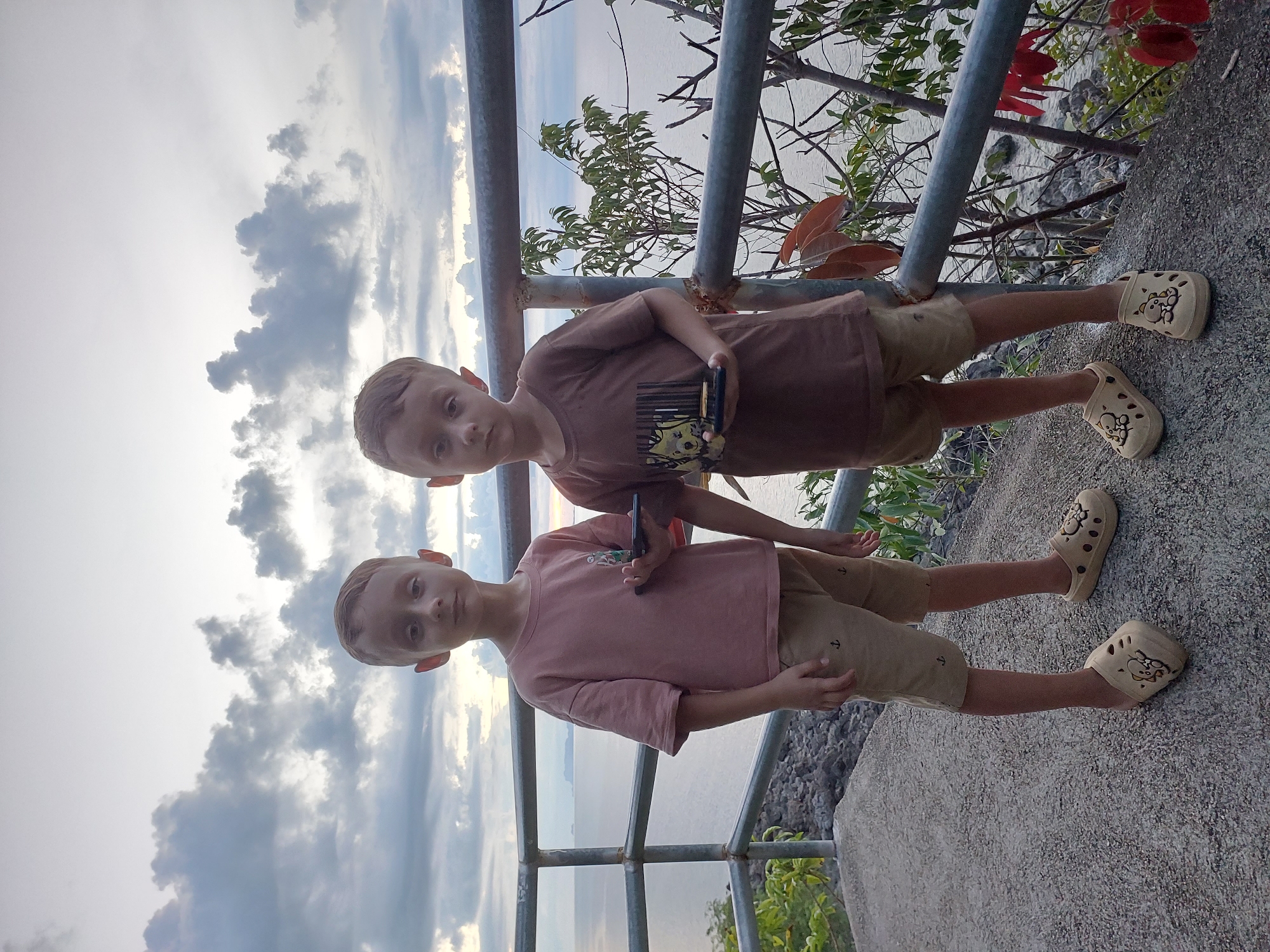 Two young boys stand side-by-side on a concrete walkway next to a metal railing, overlooking the ocean. The boy on the left wears a pink t-shirt and tan shorts, while the boy on the right wears a brown t-shirt and matching tan shorts. Both boys are wearing tan slip-on shoes with cartoon characters on them. The boy on the left holds a black smartphone in his right hand, and the boy on the right holds a black object that appears to be a small toy building structure. The sky behind them is filled with dramatic, cloudy formations, with a hint of a sunset peeking through on the horizon. The ocean stretches out before them, with distant islands visible. The location is identified as Baan Tai, Thailand, suggesting a coastal setting. The overall scene captures a moment of casual observation or play by the sea.
