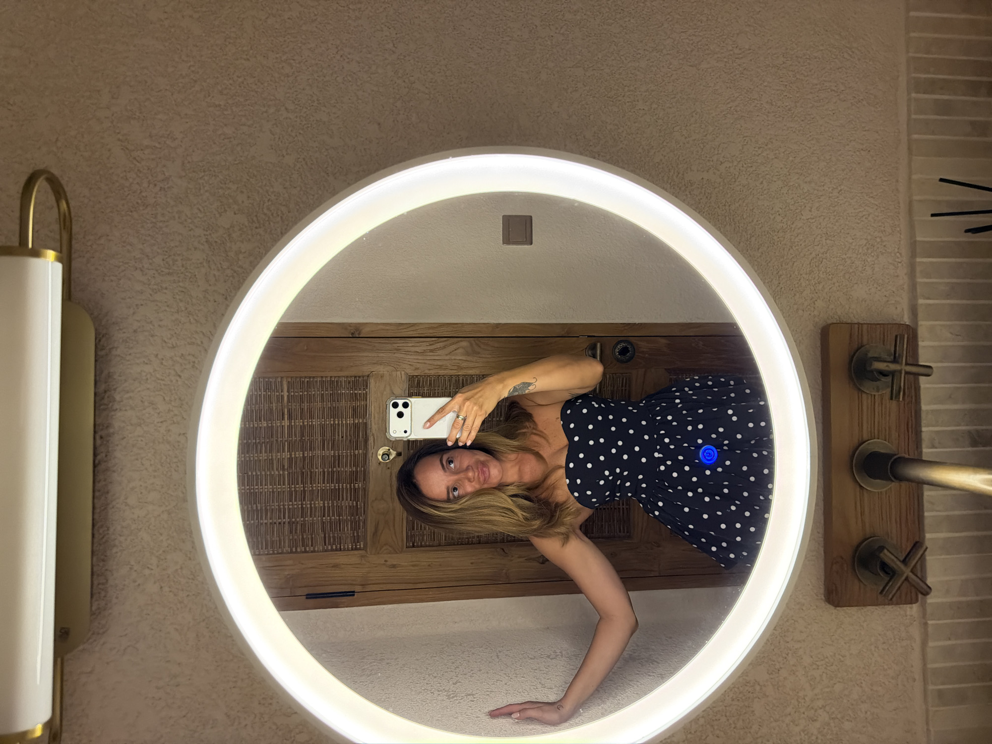 In Mengwi, Indonesia, a woman with shoulder-length brown hair, wearing a strapless navy blue dress with white polka dots, is captured in a mirror selfie. She stands with her left arm extended, touching the illuminated circular mirror. The mirror reflects a wooden door with a woven texture behind her. She holds a white smartphone in her right hand, angled to take the photo. A tattoo is visible on her left forearm. The background shows a textured wall and a wooden panel with faucets. The lighting is soft and emanating from the mirror's ring.