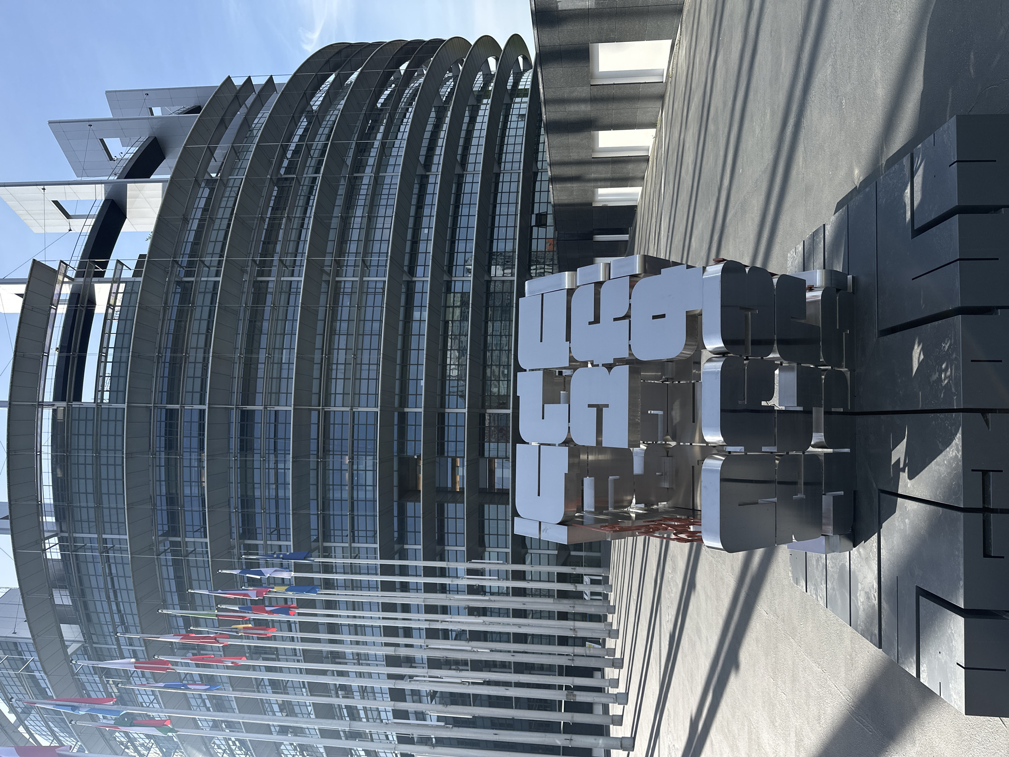 The image, taken in Strasbourg, France, showcases the modern architectural design of the European Parliament's Louise Weiss building. The building dominates the background with its distinctive curved facade, featuring numerous glass sections interspersed with grey, fin-like elements that give it a wave-like appearance.
In the foreground, a prominent metallic sculpture spells out the word "EUROPE" in large, three-dimensional silver letters, resting on a dark, block-like base. To the left, several flagpoles are visible, bearing the flags of various European Union member states, reinforcing the location's identity.
The scene is set on a bright, sunny day, characterized by a clear blue sky and sharp shadows cast by the building and the sculpture onto the paved ground, suggesting it is either late morning or early afternoon. No people are visible, allowing the focus to remain on the architecture and the symbolic sculpture in this institutional setting.