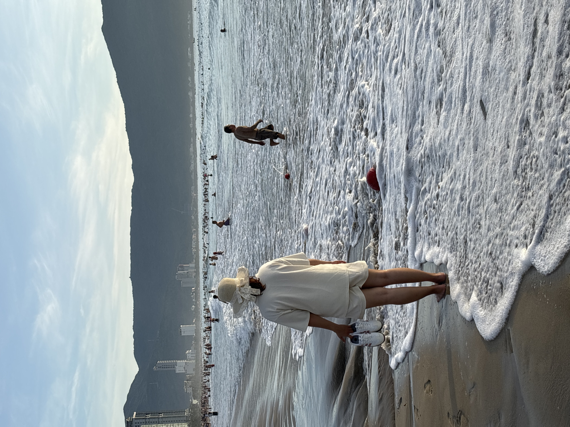 A serene beach scene unfolds in Đà Nẵng, Vietnam, with a woman in a straw hat standing on the shore, holding a pair of white sneakers. She is dressed in a matching cream-colored outfit, facing away from the camera towards the sea. The ocean is populated with numerous people swimming and enjoying the water, with a man in the mid-ground walking through the waves. In the background, the cityscape of Đà Nẵng is visible, characterized by tall buildings, with lush green mountains forming a dramatic backdrop against the sky. The sun appears to be setting, casting a warm glow over the scene, with the sky displaying soft hues of blue and white. The waves are gently lapping the shore, creating a foamy white border on the wet sand. Several red buoys are scattered in the water, marking swimming areas. The overall atmosphere is one of peaceful recreation.