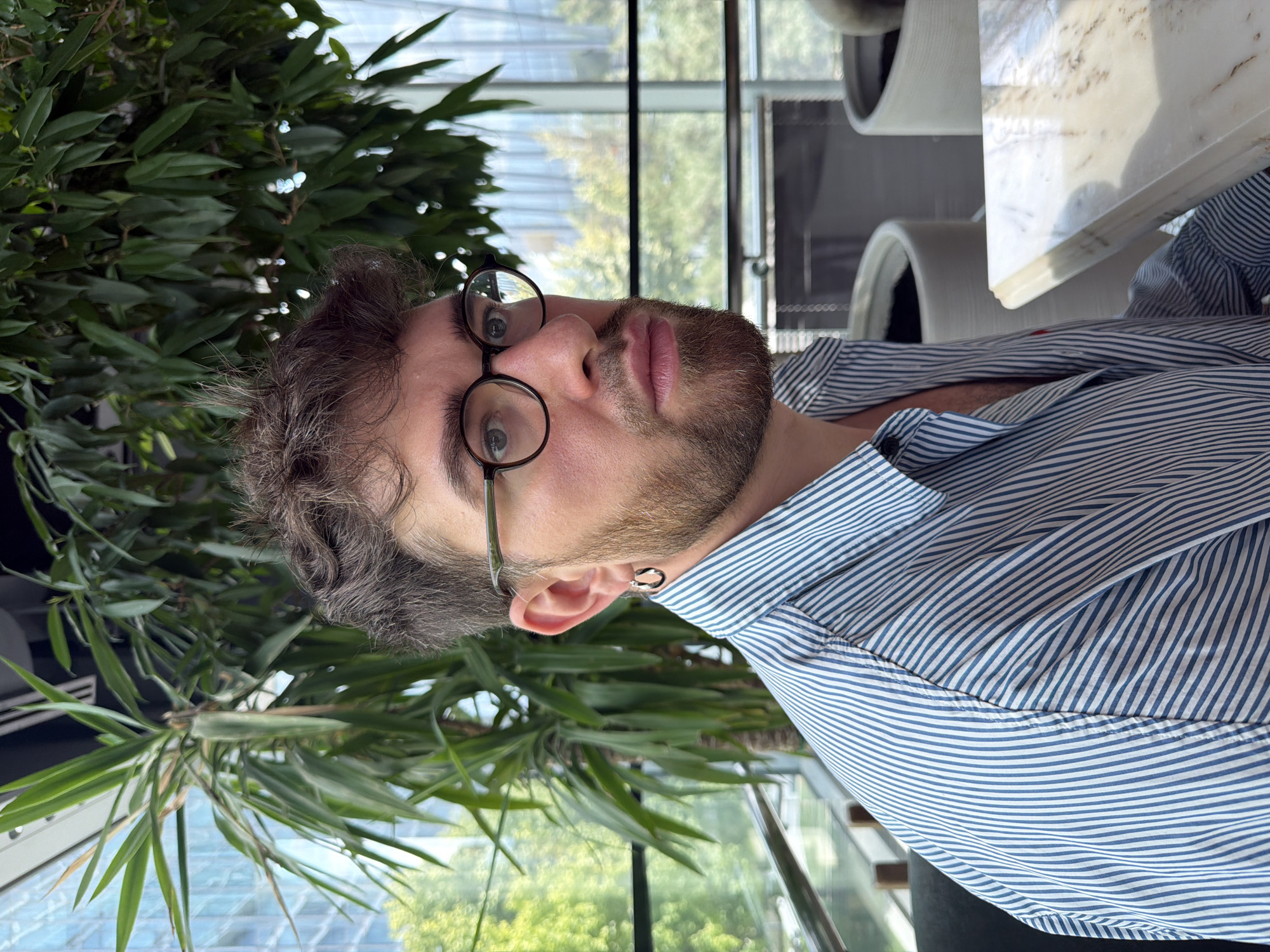A medium shot shows a young man with brown, wavy hair, a beard, and glasses, looking to his right. He is wearing a blue and white striped collared shirt. He has a hoop earring in his left ear. He is seated indoors, with large plants and a window showing trees and buildings in the background. The location is the city of Almaty, Kazakhstan.
