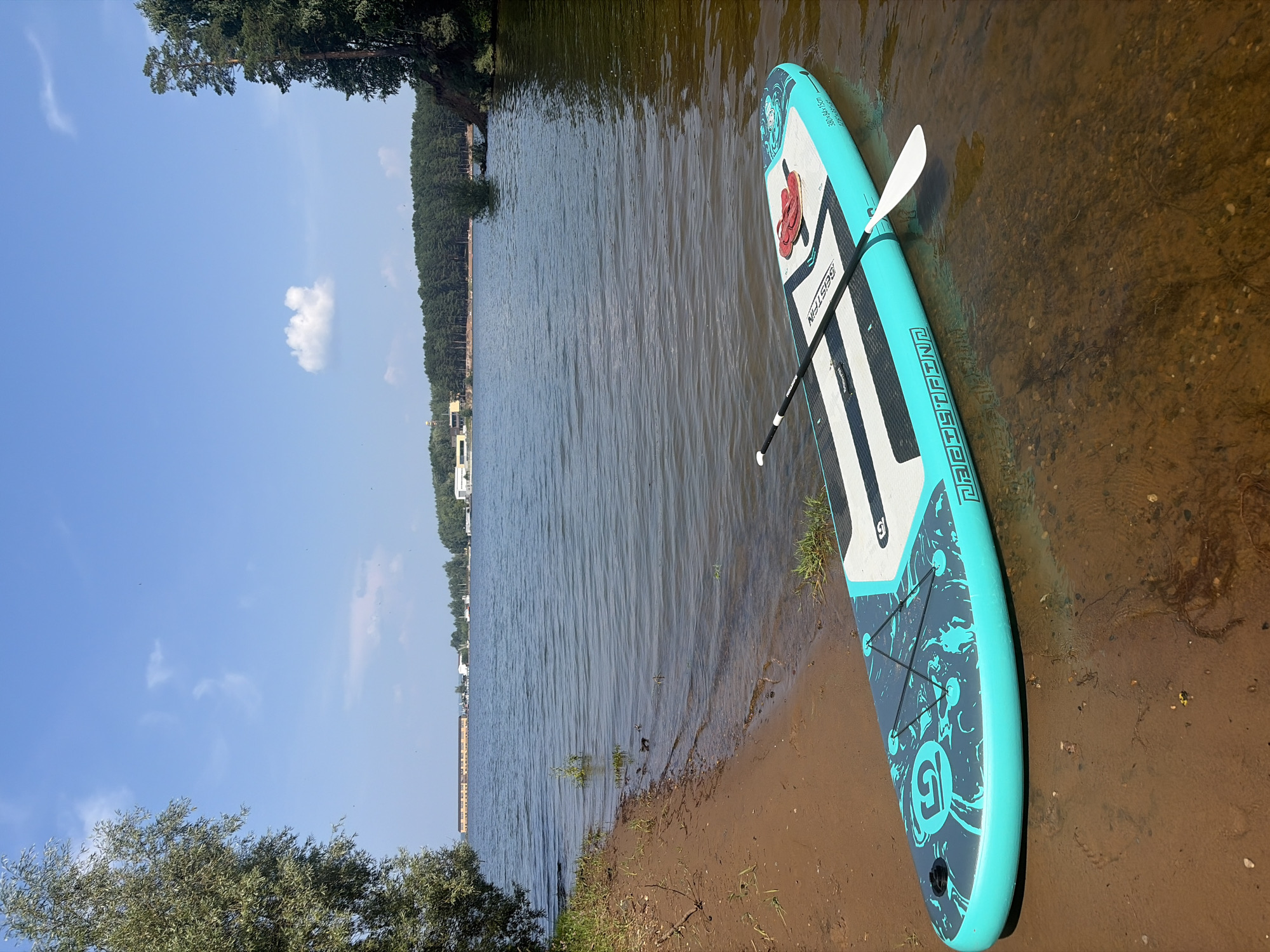 A stand-up paddleboard with a paddle and red flip-flops is partially submerged in the shallow waters of the Kama River. The sandy shore is visible in the foreground, with vegetation and trees on the left and right edges of the frame. In the background, across the river, is a tree line, a long building, and other structures, indicative of a populated area or industrial site. The sky is blue with scattered clouds. This scene is likely in the city of Chaikovsky, Russia, given the presence of the Kama River and its characteristic landscape.
