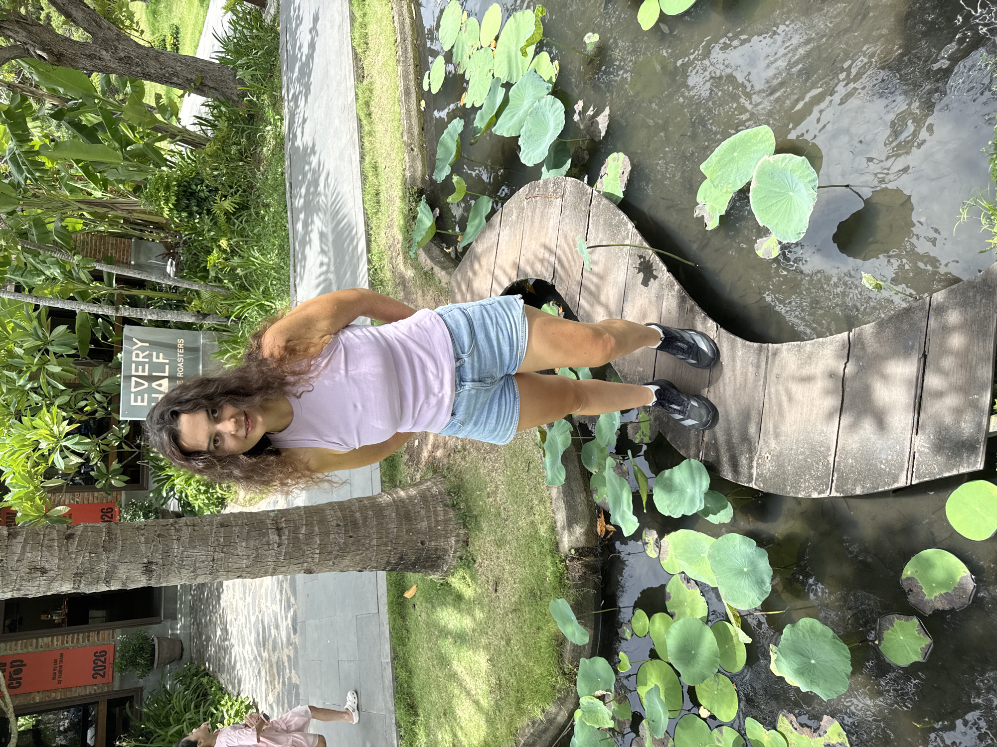 A woman with long, curly brown hair, wearing a lavender tank top and denim shorts, stands on a curved wooden walkway over a pond filled with lily pads and water. The walkway appears to be constructed from weathered wooden planks. The pond is surrounded by lush green grass and tropical foliage, including banana trees and palm trees. In the background, a building with brick accents and red banners displaying the year "2026" and Vietnamese text is visible. A sign with "EVERY HALF ROASTERS" is also present. To the left of the frame, a child in a pink dress and white clogs is partially visible, walking away from the camera. The scene suggests an outdoor setting in a city with a tropical climate, possibly a cafe or garden area in Hoi An, Vietnam, given the architectural style and vegetation.