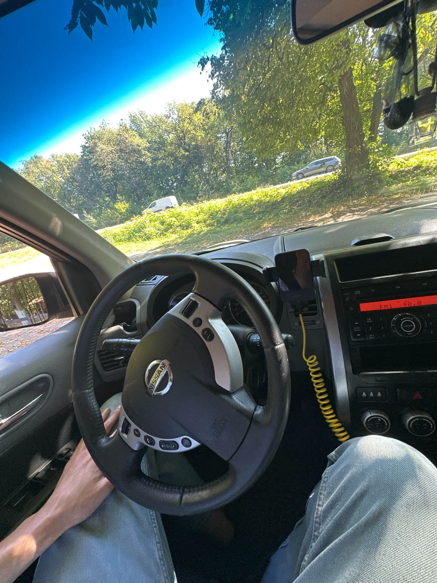 The image shows the interior of a car, a Nissan, from the driver's perspective, looking out the windshield. The driver's hands are visible on the steering wheel and on their lap, and they are wearing blue jeans. A phone is mounted on the dashboard and is plugged in with a yellow cord. The car is driving on a road lined with trees, with sunlight filtering through the leaves. In the distance, another car and a white van are visible on the side of the road. The sky is bright blue. The scene appears to be in a rural or suburban area.
