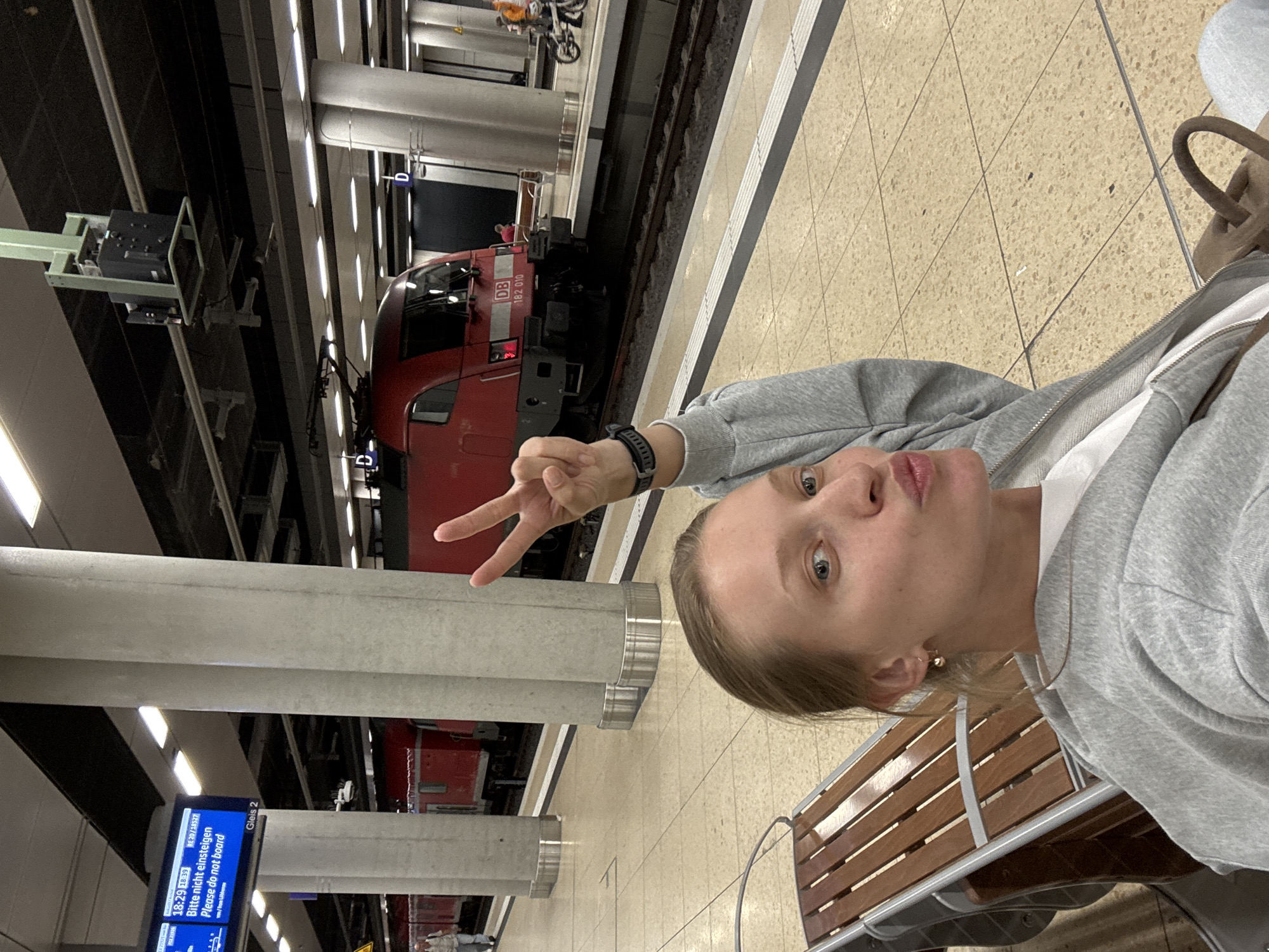 An indoor, underground train station platform is depicted, identified as being in Germany by visible signage. In the foreground, a light-skinned woman with light brown hair, pulled back, is seated on a wooden and metal bench, facing the camera. She wears a grey zippered hoodie over a white top, and small gold-colored hoop earrings. Her blue eyes are visible, and her lips are pursed in a pout. Her right hand is raised, forming a peace sign with two fingers. A dark-colored smartwatch is on her left wrist. A light brown bag is partially visible on her lap.
In the midground, a red electric locomotive, marked "DB" and "182 010", is stopped on train tracks adjacent to the platform. The platform is paved with light-colored tiles and features a white and grey striped safety line along its edge. Several thick, light grey concrete support columns are present. Overhead, a digital information screen displays "18:29 18:39" and the message "Bitte nicht einsteigen Please do not board" along with "Gleis 2" (Track 2). The ceiling is dark, with numerous linear light fixtures providing illumination, and various technical installations suspended.
In the background, to the left, a man in a light shirt walks on the platform. To the right, across the tracks, a person in an orange top stands near a bicycle, and another individual is seated further down the platform. Blue "D" signs, likely platform section markers, are visible above the tracks. The environment suggests a modern, functional train station, consistent with infrastructure found in cities such as Schönefeld, Germany.