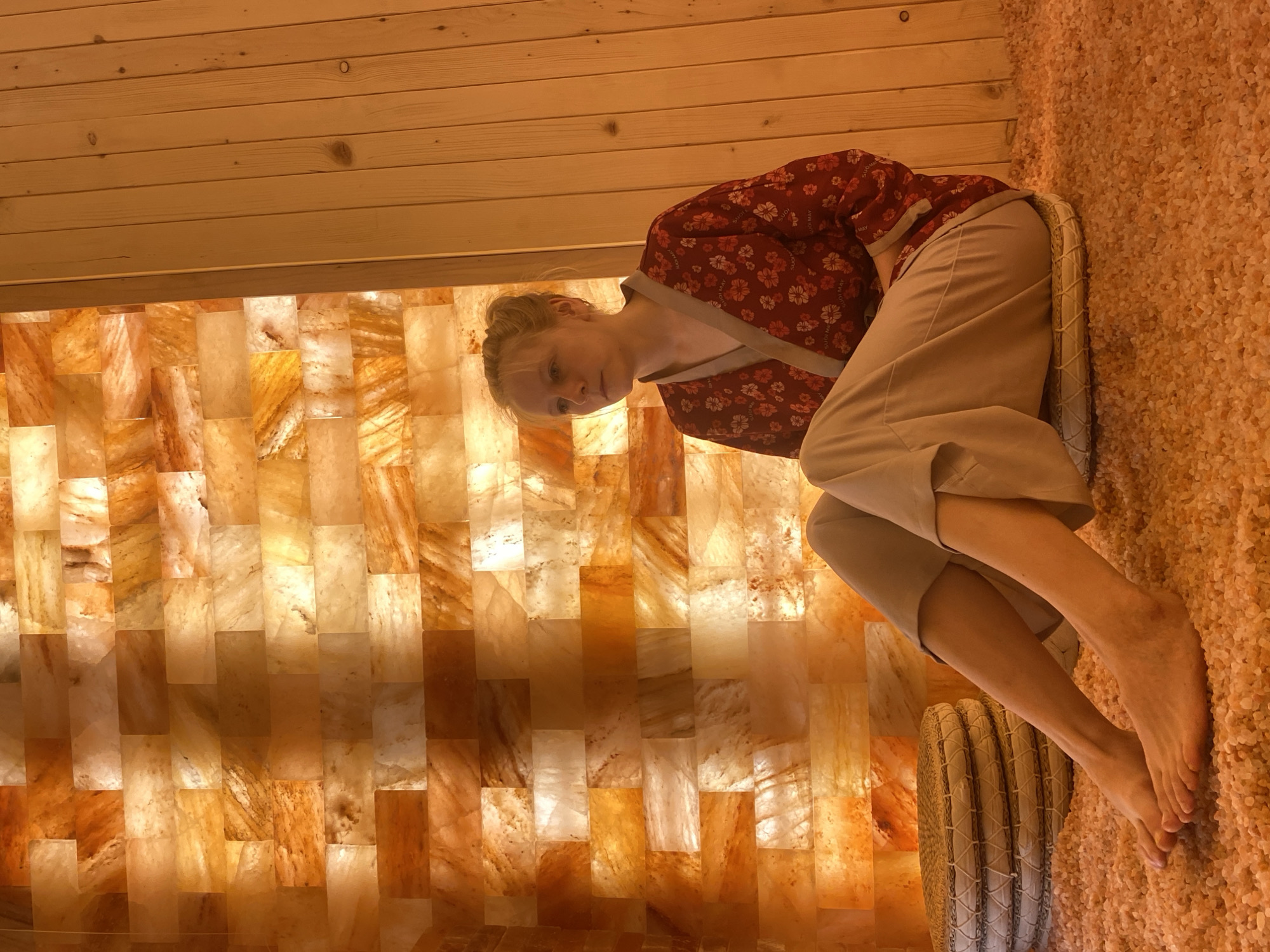 A young woman with blonde hair tied in a bun sits on a cushion on the floor of a salt room. She is wearing a red robe with a floral pattern and light-colored pants. The walls of the room are made of illuminated Himalayan salt blocks, creating a warm, orange glow. To her left are stacked cushions. To her right, the wall is made of wooden planks. The floor is covered in small, pink salt crystals. The setting appears to be a spa or wellness center in Da Lat, Vietnam.