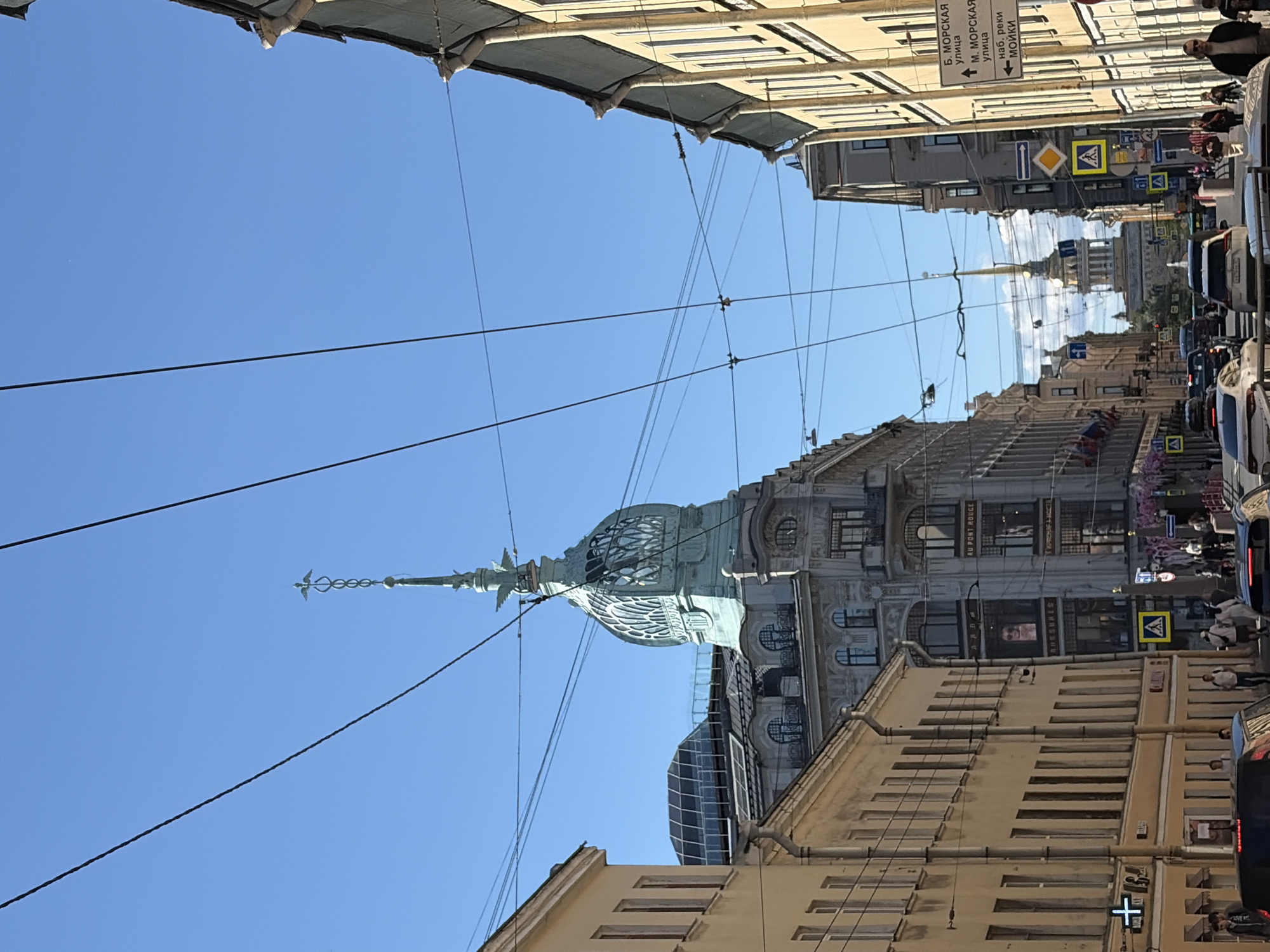 The image depicts a street scene in Saint Petersburg, Russia, likely in the city center, characterized by classical architecture and overhead wires. The perspective is looking down a street lined with tall, multi-story buildings. On the left, a light-yellow building with many windows runs along the street. On the right, a taller, more ornate gray building with decorative elements and large windows is visible, with signage that appears to be in Cyrillic. Further down the street, a distinctive building with a teal, dome-shaped roof topped by a spire with a bird-like ornament is prominent. In the distance, another tall spire can be seen. The sky is clear and blue. Several cars are present on the street, indicating it is a public thoroughfare. Pedestrians are walking along the sidewalks and crossing the street. Traffic signs and streetlights are also visible. The overall impression is of a vibrant, historic urban environment on a sunny day.