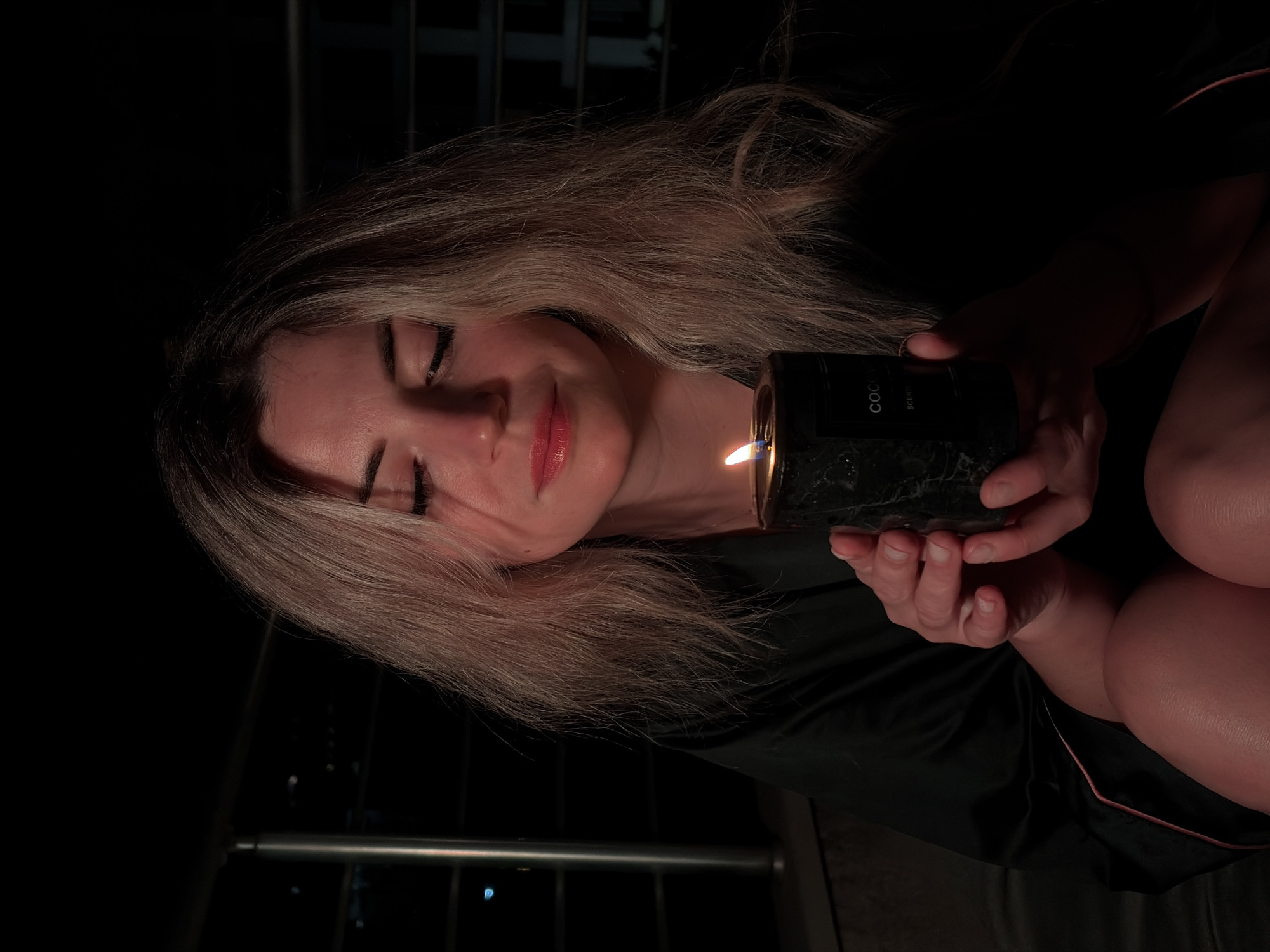 A woman with blonde hair and closed eyes holds a lit candle in her hands. She is wearing a dark robe with pink trim and her legs are visible. The background is dark, suggesting nighttime, with a balcony railing and distant city lights that could be from Budva, Montenegro. The candle is dark green marble with a label that reads "COCO SCENTED." The lighting is warm, cast by the candle, illuminating the woman's face and hands.