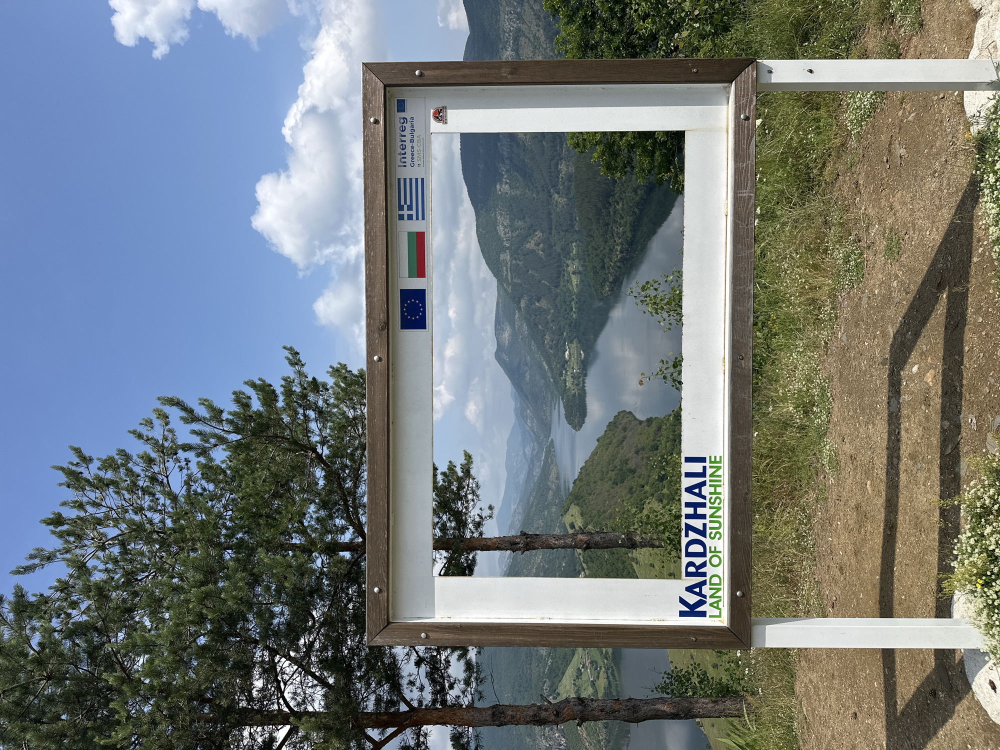 The image presents a scenic natural landscape viewed through a large, white and brown wooden sign designed as a photo frame or viewfinder. The scene is set during the daytime under a bright blue sky dotted with scattered white, puffy clouds.

Through the rectangular opening of the sign, a beautiful panorama unfolds: a wide lake or reservoir meanders through a valley surrounded by majestic, forested mountains. The water surface reflects the cloudy sky, creating a tranquil vista.

Outside the left edge of the frame, a large pine tree with dense green needles is prominently visible, its branches reaching upwards towards the sky. To the right of the sign, the ground is covered with dry grass, patches of dirt, and small white wildflowers. Long, distinct shadows are cast across the ground, suggesting bright sunlight.

The wooden frame itself displays several notable pieces of text and logos. Vertically aligned on the right side of the frame, in large blue letters, is "KARDZHALI", with "LAND OF SUNSHINE" written in smaller green letters below it. On the top left portion of the frame, the "Interreg Greece-Bulgaria" logo is visible, alongside the national flags of Greece and Bulgaria, and the flag of the European Union, indicating a cross-border initiative. The setting is Star Chitak, Bulgaria.