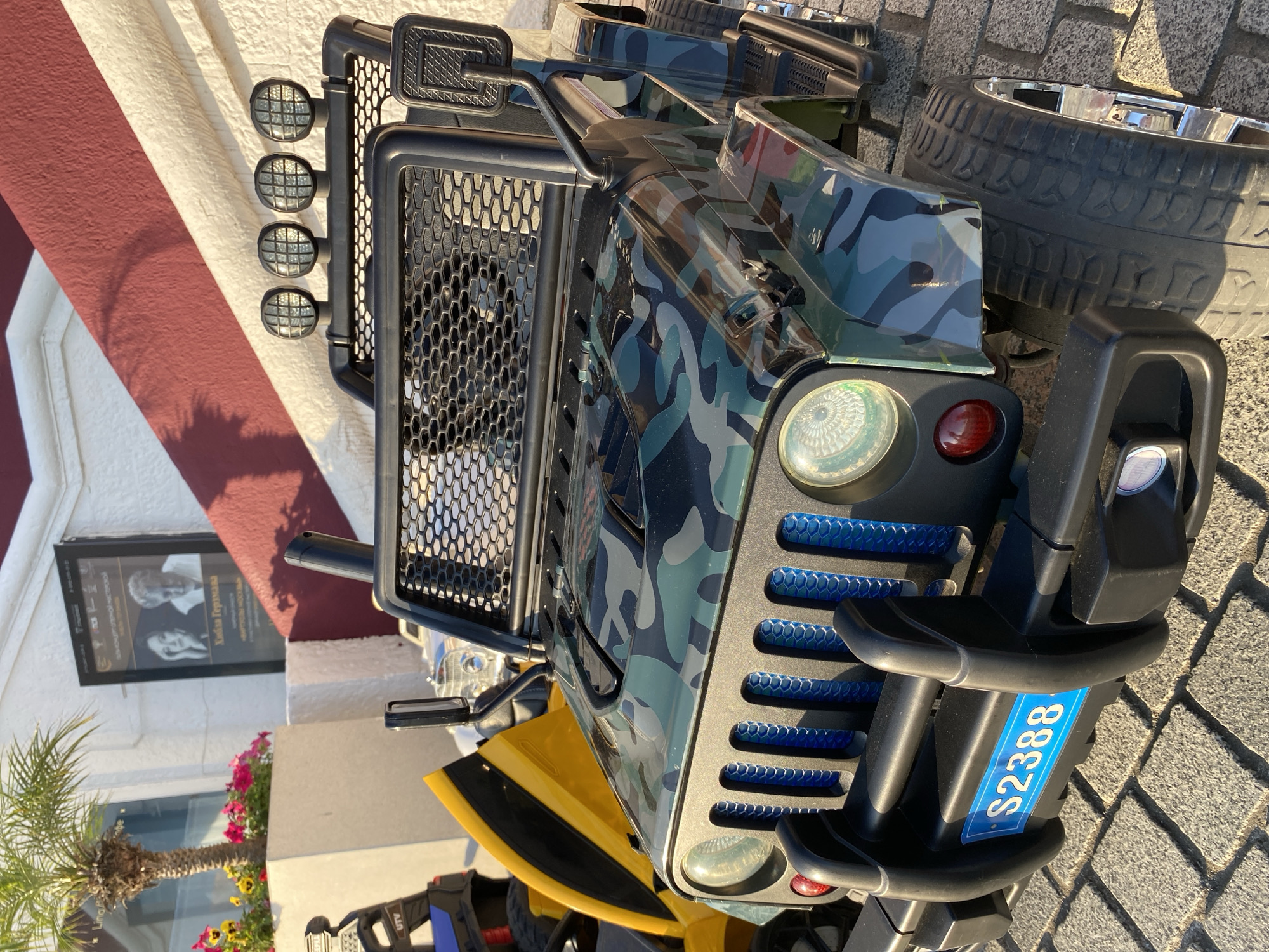 This media file shows a close-up, rotated view of a child's toy jeep parked on a paved surface, likely outdoors. The jeep is predominantly green and black with a camouflage pattern, and features a black grille with blue accents, four round headlights on the front, and a red taillight. A blue license plate with white text "S2388" is visible on the front bumper. The jeep has a large, chrome-rimmed tire and a black side mirror. In the background, a building with a red and white striped facade is visible, along with a framed poster containing images of two people and Russian text. A small palm tree and some pink and yellow flowers are also present, suggesting a warm climate. The lighting indicates it is daytime, possibly late afternoon or early morning, given the shadows cast on the building. The overall scene suggests a residential or recreational area, possibly near a vacation spot.