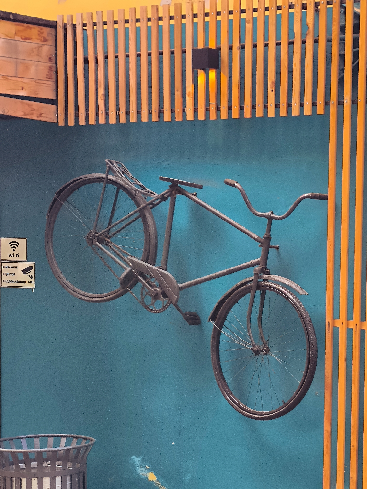 The image shows a dark-colored vintage bicycle mounted on a teal-colored wall. The bicycle is positioned diagonally with its front wheel pointing towards the lower right and the rear wheel towards the upper left. The bicycle has a rack over the rear wheel. To the left of the bicycle, there are two signs mounted on the wall. The upper sign has a Wi-Fi symbol and the text "Wi-Fi". The lower sign has a CCTV camera icon and text in Cyrillic that appears to indicate video surveillance. Above the bicycle and signs, there is a structure made of vertical wooden slats painted orange, with some wooden panels to the left. A black light fixture is visible among the wooden slats. In the lower left corner, a metal trash can is partially visible. The overall scene appears to be an indoor or covered outdoor area, possibly a public space like a cafe or a street art installation. The location is identified as the city of Karaganda, Kazakhstan.