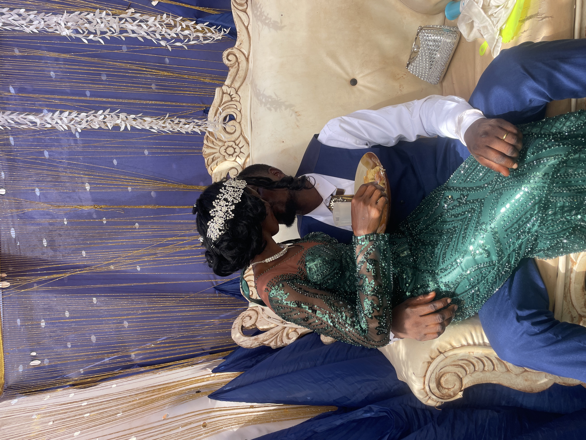 A horizontal eye-level shot depicts a couple in formal attire seated on an ornate off-white couch with a navy blue draped cloth at the base, set against a backdrop of vertical blue and gold tinsel adorned with white leafy garlands, potentially within Ogbe, Nigeria. The woman, on the left, wears a green, long-sleeved, sequined dress and a jeweled headpiece. The man, on the right, is in a navy suit and white shirt. They are face-to-face, sharing food from a golden plate held by the man. The man wears a wedding band on his left hand. Next to the couple, on the couch, are a silver clutch and a few indiscernible objects wrapped in white fabric.