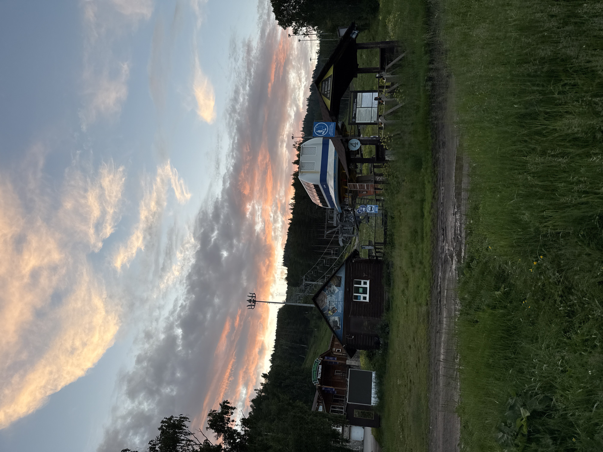 The image depicts a scenic view of a ski resort in Syrostan, Russia, at sunset. The sky is filled with dramatic clouds painted in hues of orange and pink, contrasting with the soft blue of the upper atmosphere. In the foreground, lush green grass covers the ground, with a muddy track running through it. Several wooden structures are visible, including a small cabin with a white-framed window and a larger building with a sign that reads "Sunny Valley Restaurant".
Dominating the middle ground is a ski lift with a white and blue gondola station. The ski lift extends up a forested hill, disappearing into the trees. A ski lift tower stands tall, and other pylons are visible along the slope. Signs with pictograms and Russian text are posted near the gondola station. The overall environment suggests a recreational area surrounded by nature, with the setting sun casting a warm glow over the landscape. There are no people visible in the image.