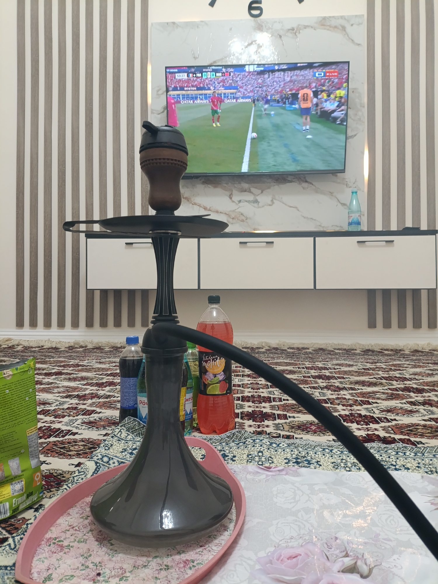 A low-angle shot shows a hookah in the foreground, placed on a pink oval tray with a floral pattern. The hookah is dark gray with a black hose. Behind the hookah, on a patterned rug, are several bottles of beverages, including a large red bottle with a label featuring fruit, a blue bottle, and other smaller bottles. A green package is visible to the left of the hookah. In the background, a wall with vertical stripes of white and light wood panels is present. A black and white television screen displays a soccer match, with players in red and white uniforms visible. A white cabinet with two drawers is positioned below the television. A large clock with a black frame and large numbers is mounted on the wall above the television. The lighting in the room appears to be artificial, with a soft glow emanating from the television and possibly other light sources not visible in the frame. The overall scene suggests a casual setting, possibly a living room in Ashgabat, Turkmenistan, where someone is watching television and enjoying a hookah.