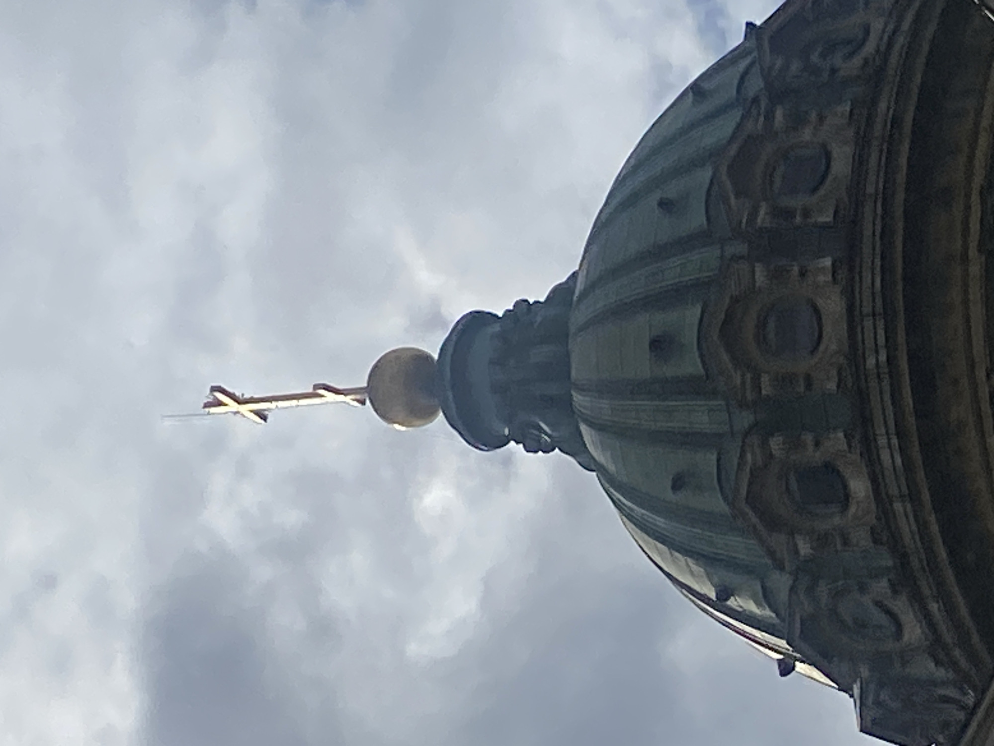 The image shows the top of a dome, likely part of a church or cathedral, under a cloudy sky. A golden cross sits atop a golden orb, which is itself mounted on a dark, ornate structure that is part of the dome's apex. The dome itself is a deep green and appears to be made of metal, with visible architectural details such as ribbed sections and decorative molding with circular openings. The overall impression is one of grandeur and religious architecture. This structure is consistent with the appearance of the dome of Saint Isaac's Cathedral in Saint Petersburg, Russia, which is a prominent landmark.