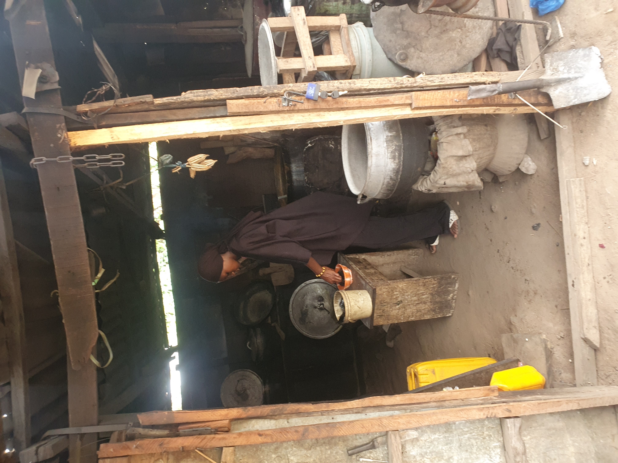 The image depicts a rustic, semi-enclosed cooking or work area, likely in Lokoja, Nigeria, with a single person engaged in food preparation. The setting is characterized by a dirt floor and a simple structure built from rough wooden beams and planks. Natural light streams in from openings on the left side, indicating daytime.
In the center of the frame, a person, dressed in a dark brown or black long-sleeved top, trousers, and a head covering, is bent over a large, dark metal pot. They hold a small orange bowl in their right hand, which is adorned with several gold-colored bangles. Multiple other dark metal pots of various sizes are visible on the ground around the person, with some appearing to be on low stands. Faint smoke or steam rises from the pots, suggesting active cooking. A small, light-colored bucket or pot sits on a wooden stool beside the person.
The immediate surroundings are cluttered with various utilitarian objects. To the right, a large spade or shovel rests on a wooden beam. A blue padlock with a visible "32GB" inscription and a key is also placed on a wooden beam above the cooking area. Further right, large, round concrete or stone objects are visible on the ground. On the far left, metal chains are attached to a vertical wooden post, and what appears to be decorative dried plant material hangs from a beam. Two bright yellow plastic jerrycans are on the ground towards the bottom right. The overall impression is that of a functional, basic domestic or commercial cooking space.