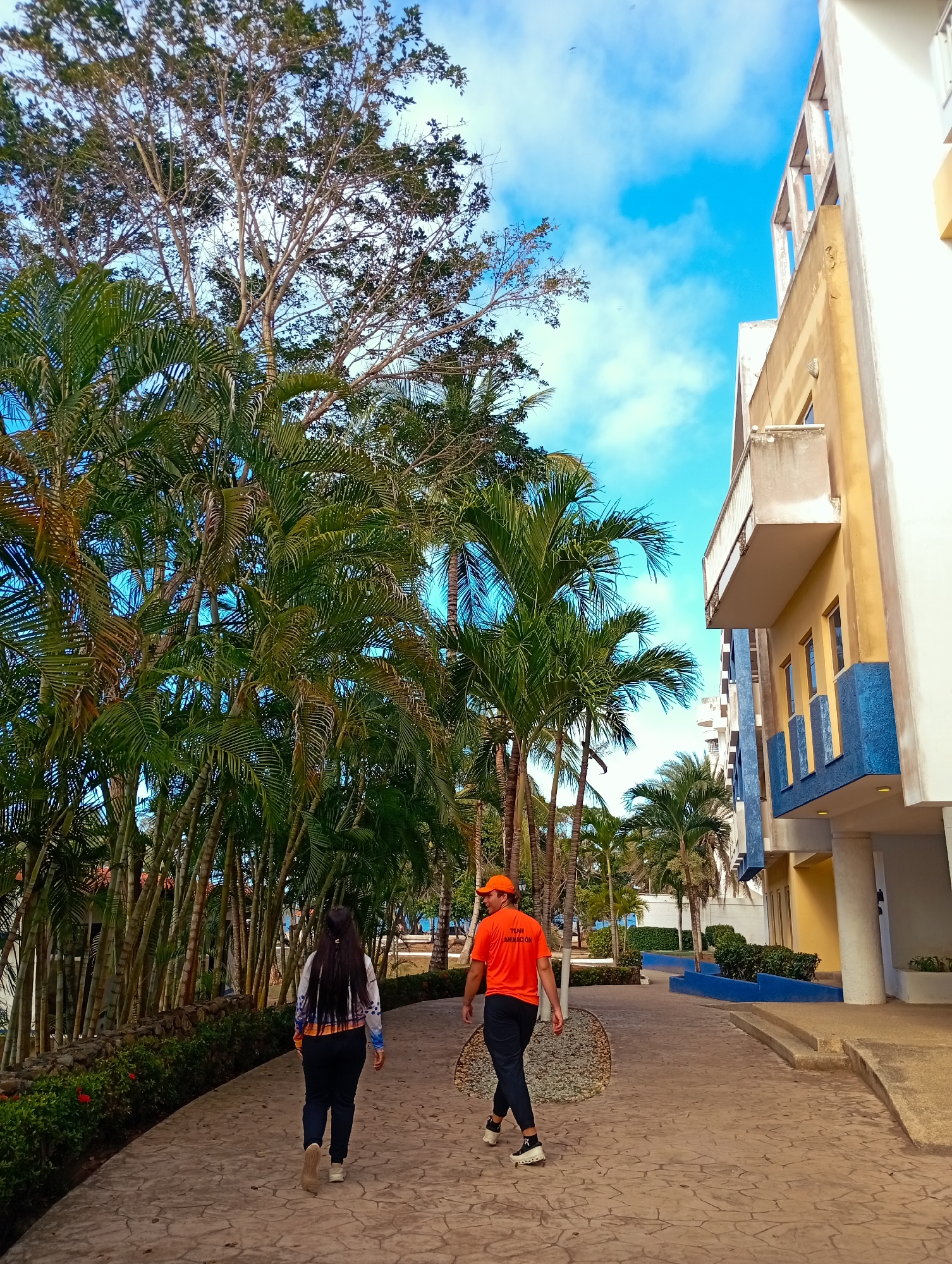 Two people are walking on a stone path in Playa El Agua, Venezuela. The person on the left is a woman with long dark hair, wearing a patterned white and blue shirt, dark pants, and light-colored shoes. The person on the right is a man wearing an orange T-shirt with the words "TEAM ANIMACION" on it, dark pants, a matching orange hat, and white sneakers. The path is lined with lush tropical vegetation, including palm trees and flowering bushes. To the right of the path, there is a yellow and blue building. The sky is blue with some clouds visible above the trees and buildings.