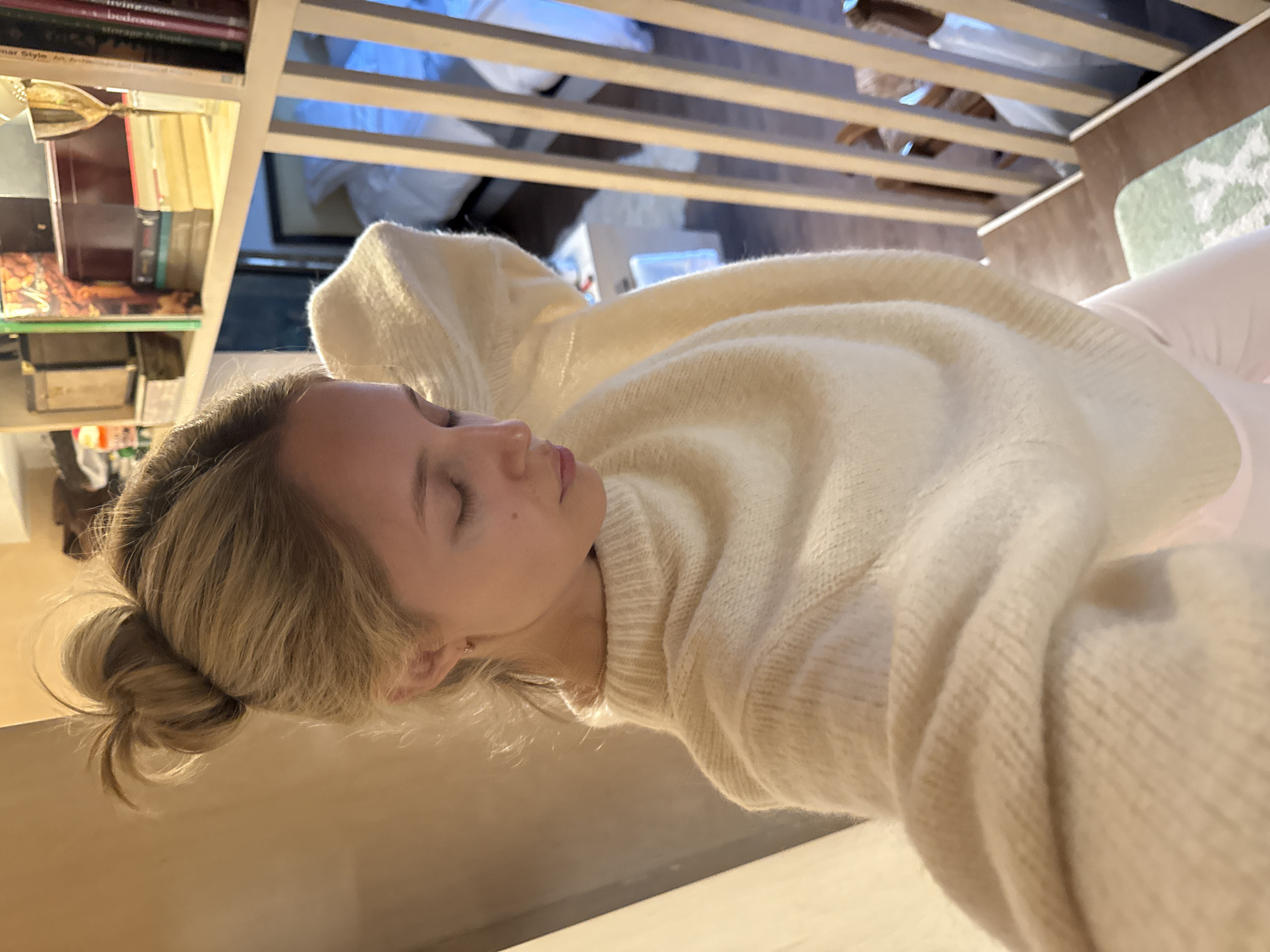 A young woman with blonde hair pulled into a messy bun is looking down with her eyes closed. She is wearing a fluffy white sweater and pink leggings. She is standing on a wooden staircase. In the background, there are bookshelves filled with books and decorative items, and a glimpse of a bedroom with a bed.