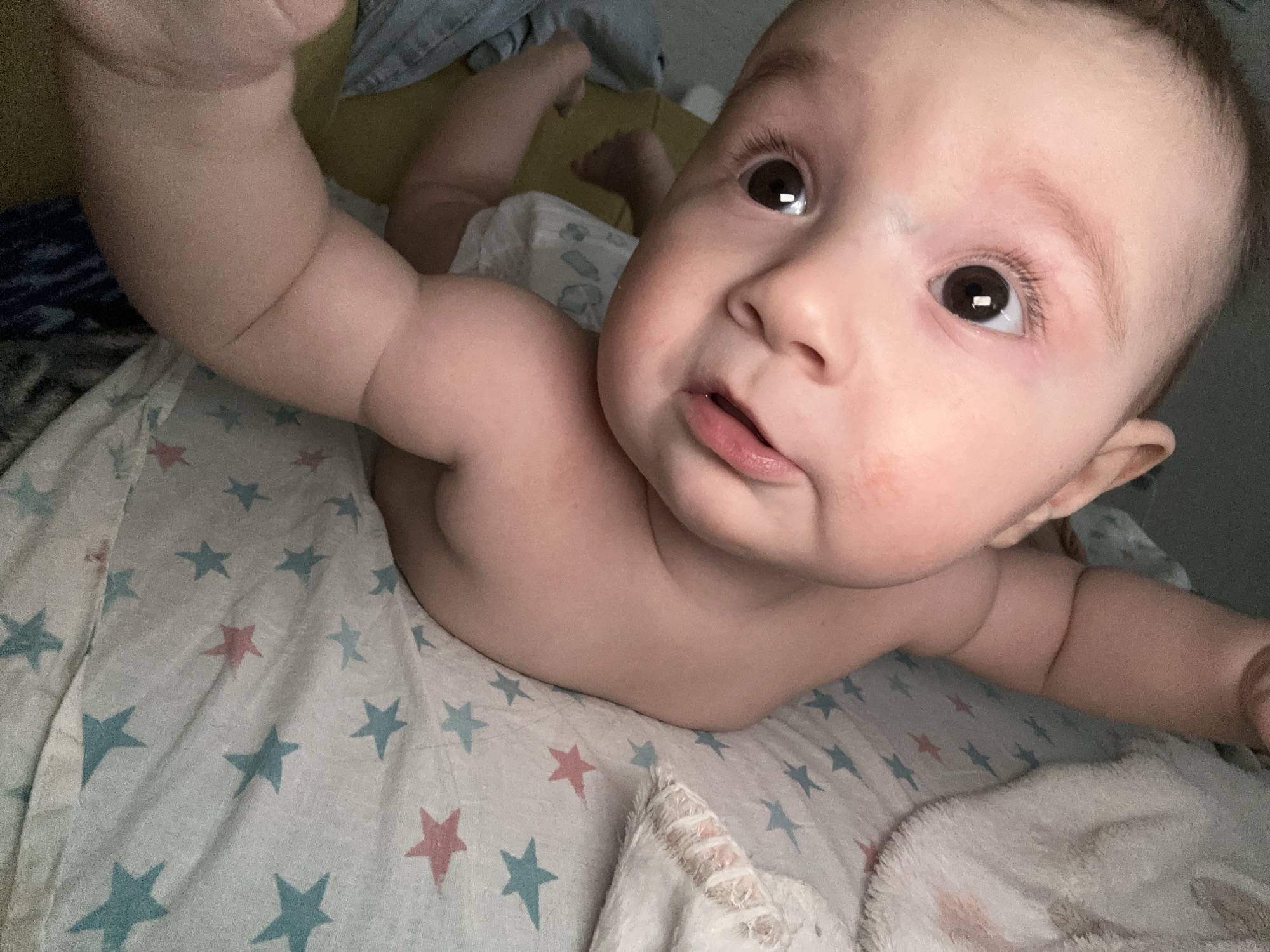 A baby is lying on its stomach on a blanket decorated with red and blue stars. The baby is facing upwards with eyes open and mouth slightly ajar. One arm is raised and bent. The location is Chaikovsky, Russia.