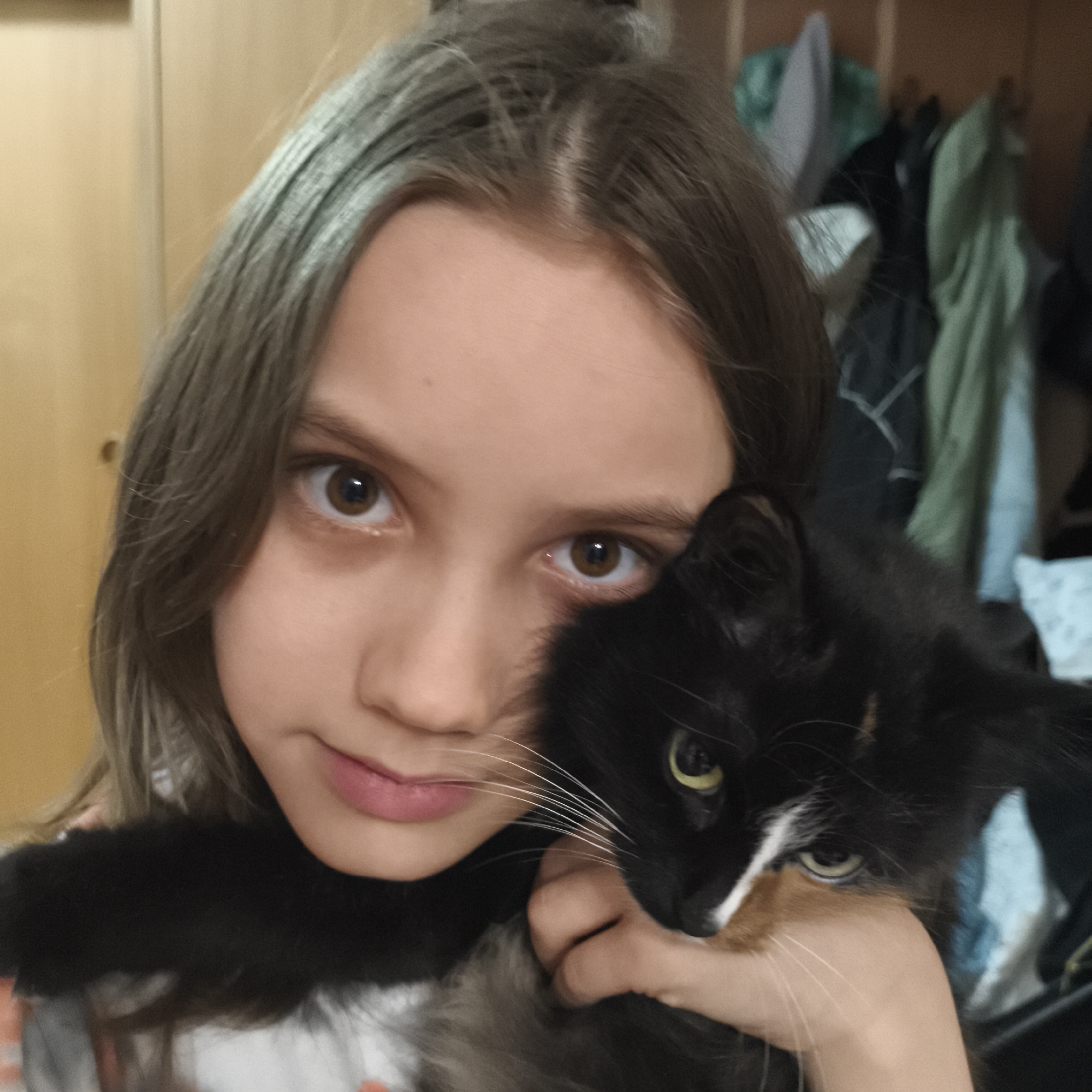 A young person with brown hair is holding a calico cat. The person's face is close to the cat's, almost touching. The cat has black fur with patches of orange and white. The background appears to be a closet with clothes hanging. The location in the city of Chaikovsky, Russia, is unconfirmed based on the provided image.