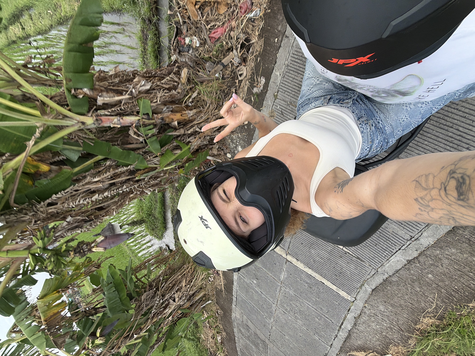 A woman, wearing a light yellow motorcycle helmet and a white tank top with blue patterned jeans, is taking a selfie on a motorcycle. She is winking and holding up two fingers in a peace sign. The motorcycle is on a paved road with green rice paddies and banana trees in the background. The location appears to be in a rural area outside of North Kuta, Indonesia.