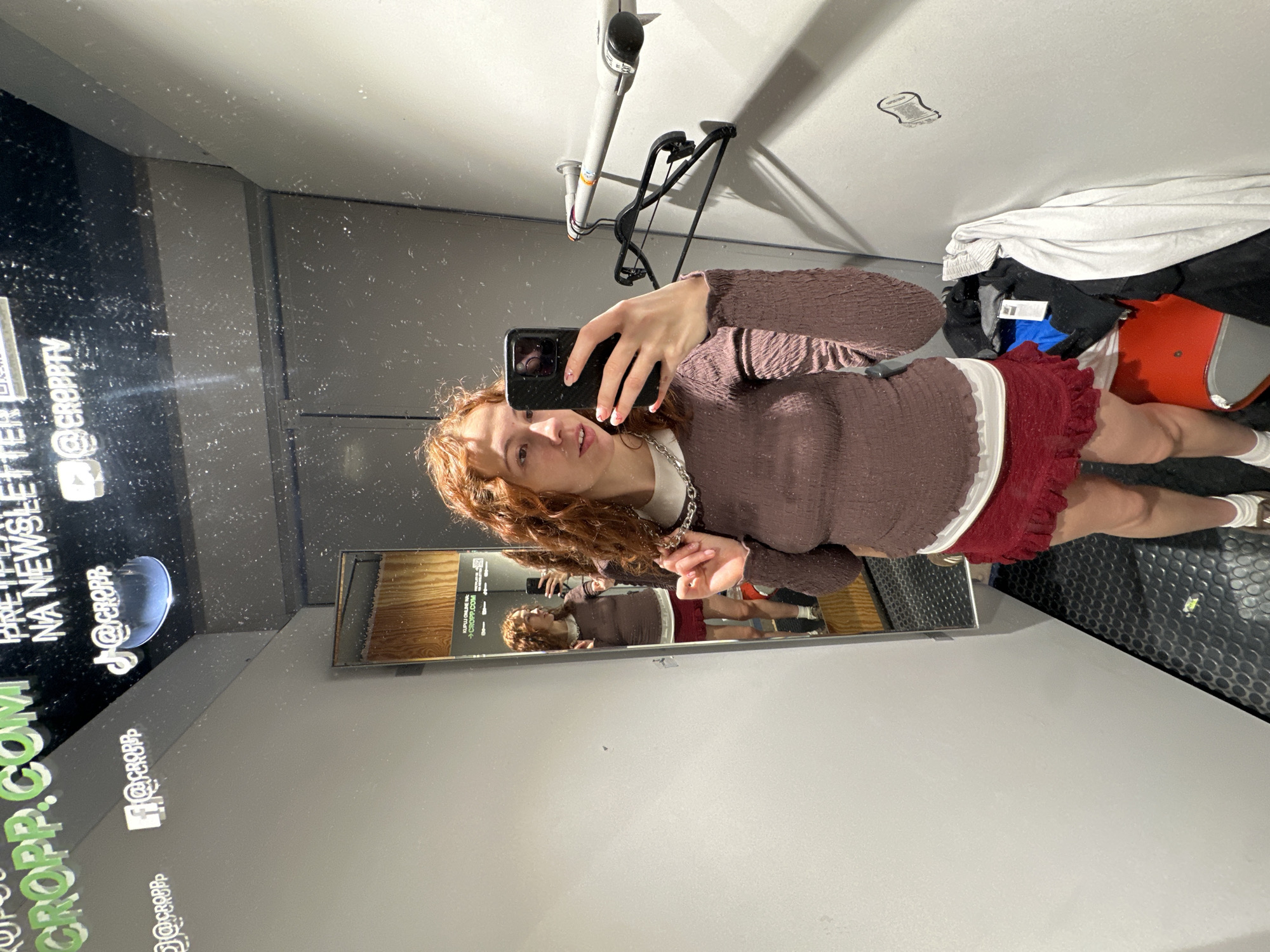 A young woman with red curly hair is taking a selfie in a dressing room. She is wearing a long-sleeved, textured purple top, a white undershirt, and a short, ruffled red skirt. A silver chain necklace is visible around her neck. She is holding her phone in her right hand, and her reflection can be seen in a mirror on the left.
The dressing room is cramped, with gray walls and a black rubberized floor. Clothing items are piled on an orange and gray stool in the corner. A clothing rack is visible in the upper portion of the image. The location appears to be a retail space, based on signage on the wall including "@CROPP.TV", "@CROPP", and the word "CROPP". The precise location in Novi Sad, Serbia is not discernible from the image.