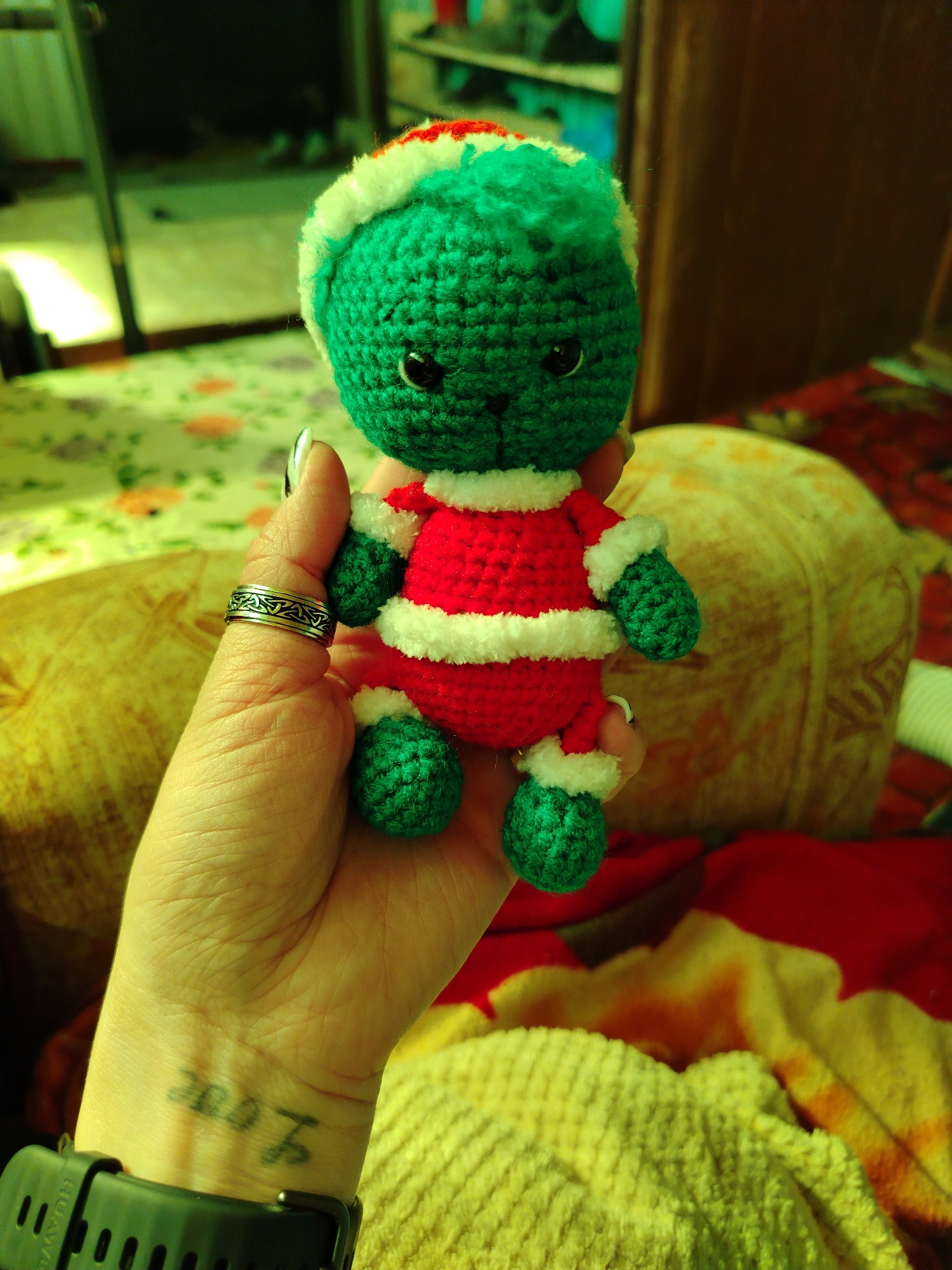 A hand holds a small, crocheted green, red, and white doll. The doll has a green head and limbs, a red body, and white trim. The hand has pale skin, a silver ring with a repeating pattern on the ring finger, a black smartwatch on the wrist, and a faded "Love" tattoo on the wrist. The background is blurry and indistinct but appears to be a domestic interior. The image doesn't contain information about the location in the city of Гожан, Russia.
