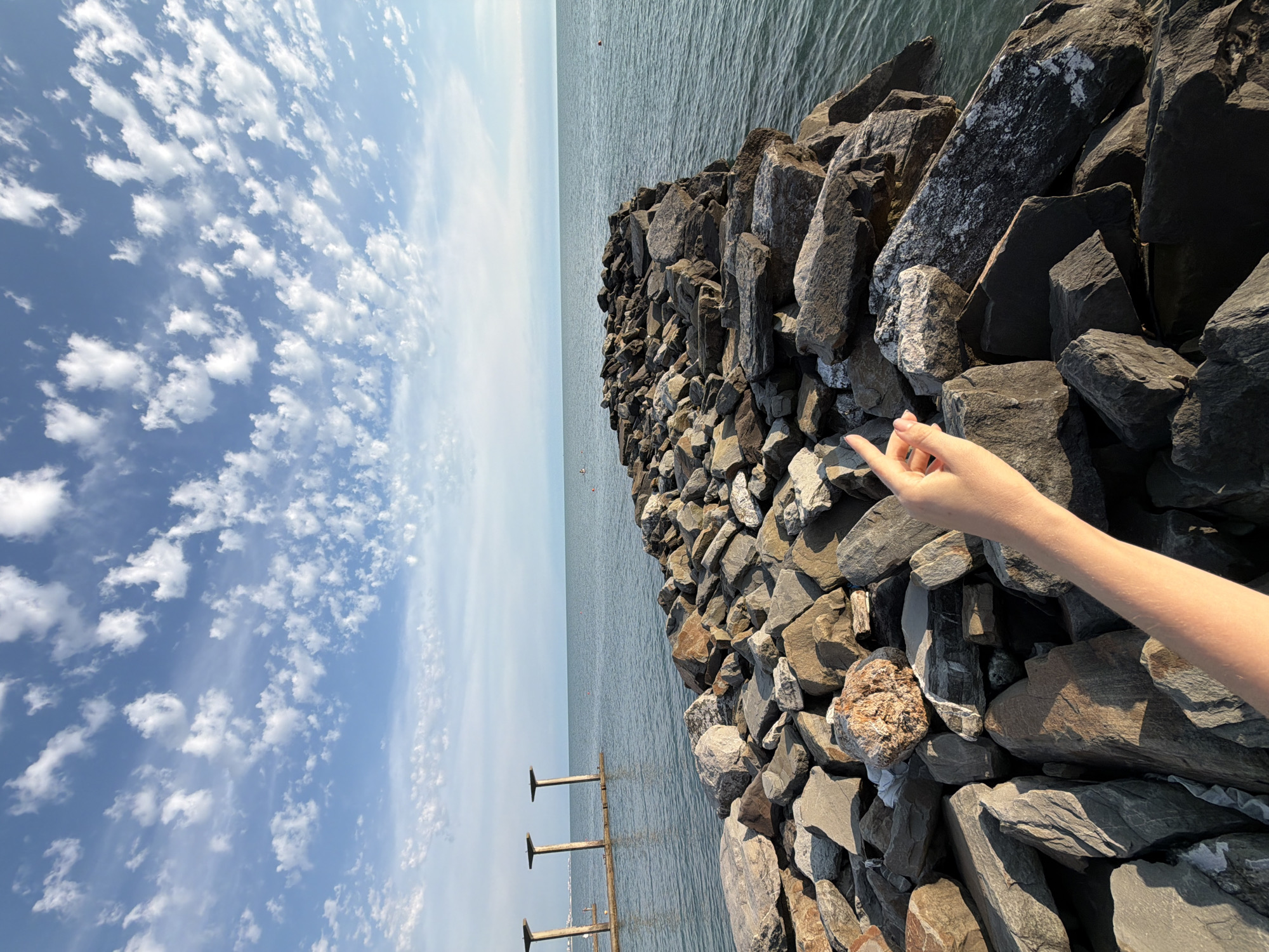 The image depicts a rocky pier extending into a calm body of water under a sky with scattered white clouds. In the foreground, a person's arm and hand are visible, with the index finger pointing towards the end of the pier. The pier is constructed from large, irregular grey and brown stones. In the background, the water stretches to the horizon, meeting a pale blue sky. There are three vertical structures with horizontal beams on a wooden platform in the water to the left of the frame. A few small buoys are visible in the water. The overall scene suggests a serene outdoor environment, likely a coastal area.