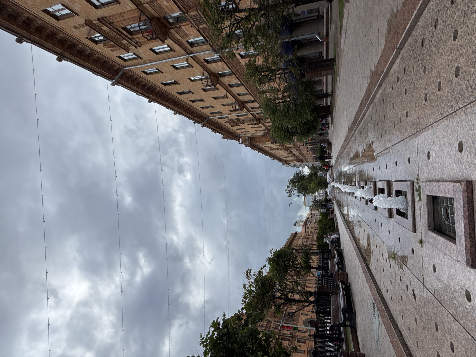 A long, narrow street with fountains running down the center is pictured on a cloudy day. The street is lined with tall, brick buildings on both sides. The buildings have many windows and balconies. There are trees planted along the sidewalk. People are walking on the sidewalk and sitting on benches. The fountains are in a long, rectangular pool with water jets shooting up. The sky is overcast and gray.