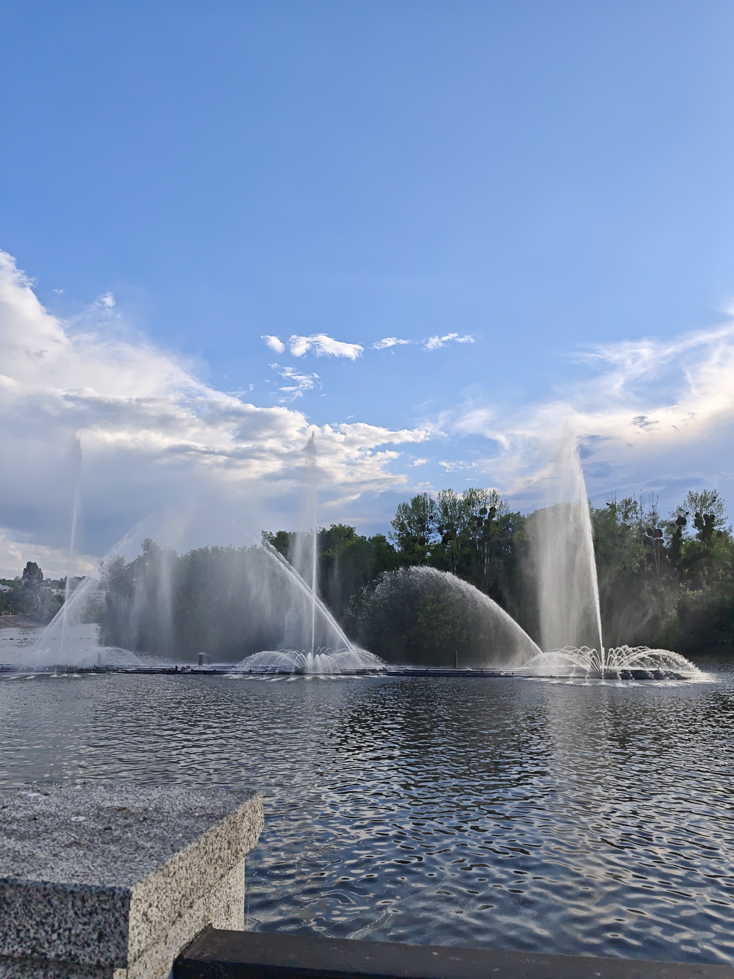 This media file captures a scenic view of a large fountain display on a body of water, likely a river or lake, with a lush green treeline in the background. The sky above is a vibrant blue, adorned with scattered white clouds, suggesting a pleasant day. The time of day appears to be late afternoon or early evening, indicated by the soft lighting and the angle of the sunlight.
The fountain is the primary subject, with multiple jets of water creating dramatic arcs and sprays. Some jets are tall and powerful, reaching high into the air, while others form wider, fan-like shapes closer to the water's surface. The water itself reflects the sky and the surrounding greenery, adding depth and shimmer to the scene.
In the foreground, a stone ledge or embankment is visible, possibly part of a promenade or viewing area. The rough texture of the stone contrasts with the smooth, rippling surface of the water. The overall atmosphere is one of tranquility and natural beauty, with the dynamic movement of the water providing a focal point.
The location is identified as Vinnytsia, Ukraine. There are no visible people or text within the frame. The weather appears to be fair and partly cloudy. The scene evokes a sense of peaceful enjoyment of a public space.