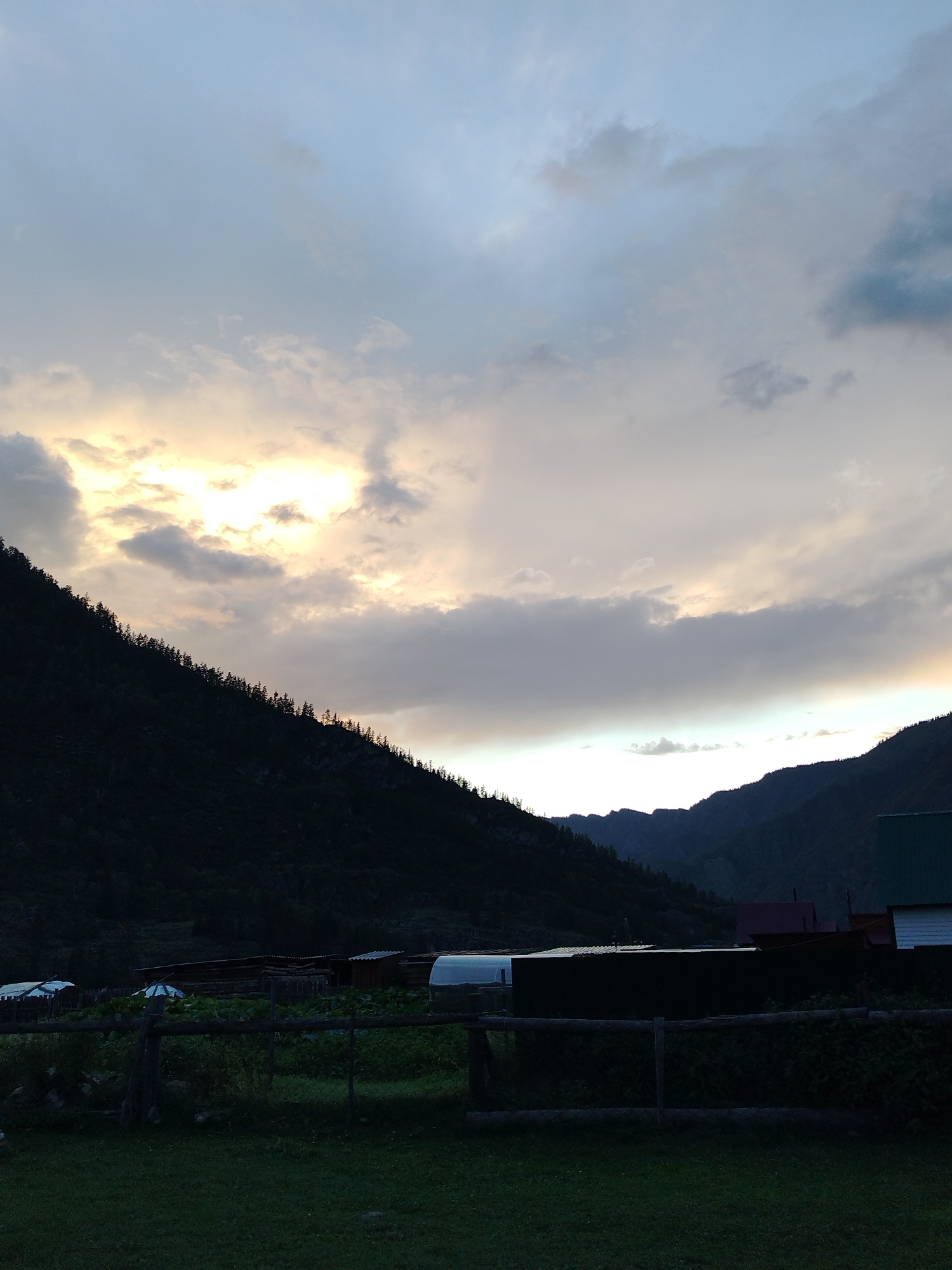 The image depicts a rural landscape at dusk, likely in the city of Iodro, Russia. The sky is filled with dramatic clouds, with a bright area of sunlight breaking through on the left side, casting a warm glow. A steep, tree-covered mountainside dominates the left half of the frame, silhouetted against the sky. In the foreground and midground, there are several low-lying buildings, possibly farm structures or houses, with various roofs visible. A wooden fence runs across the lower portion of the image, separating a grassy area from the structures behind it. The overall impression is one of natural beauty and a tranquil, remote setting as daylight fades.
