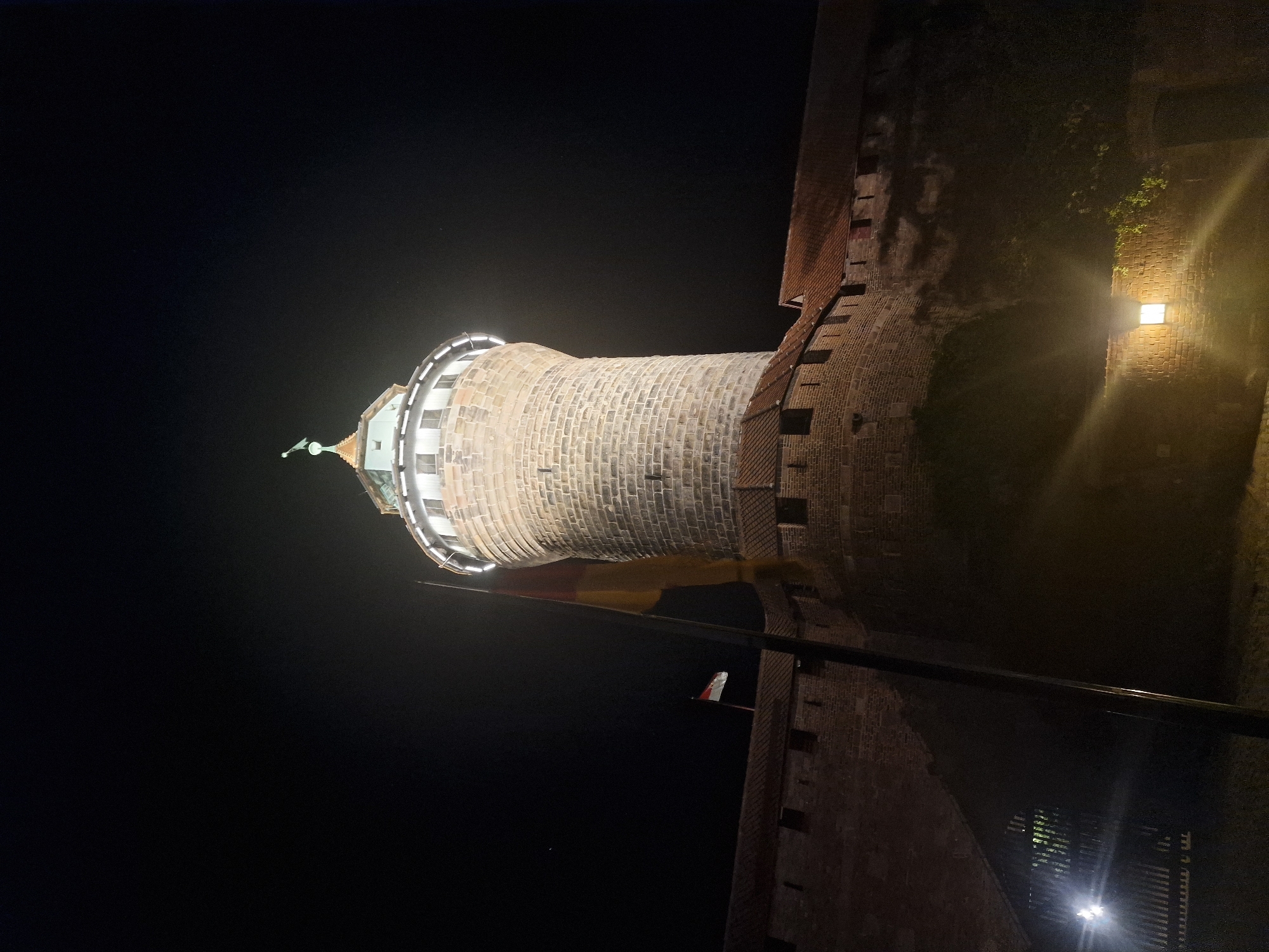 The image depicts a nighttime view of the Nuremberg Castle in Nuremberg, Germany. The castle features a prominent, circular stone tower illuminated from within, with a green spire at its apex. A German flag, with its black, red, and gold bands, is visible on a flagpole in front of the tower, alongside a smaller flag. The castle walls, constructed of stone and brick, are visible in the foreground and extend across the frame. The scene is lit by artificial lights, casting a warm glow on the architecture and creating lens flares in the foreground. The sky is dark and clear, with a few distant lights visible. The overall impression is one of historical grandeur and nighttime illumination.