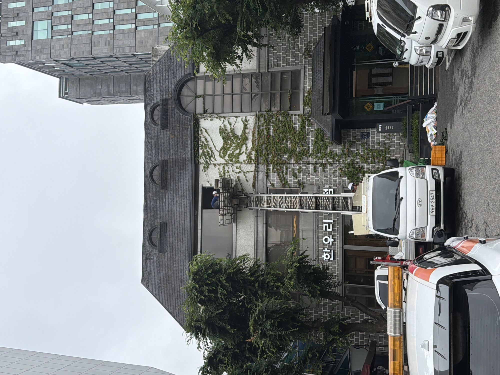 An exterior view of a building in Seoul, South Korea, shows a delivery or moving operation in progress. A ladder lift extends from a white truck to an upper window of a grey stone building, where a person in blue work clothes is visible on the lift platform. The building has a dark, sloped roof with dormer windows, and ivy climbs the facade. A white truck with an orange light bar is parked in the foreground, and another white truck is positioned behind it. The scene appears to be on a street with trees lining the sidewalk.